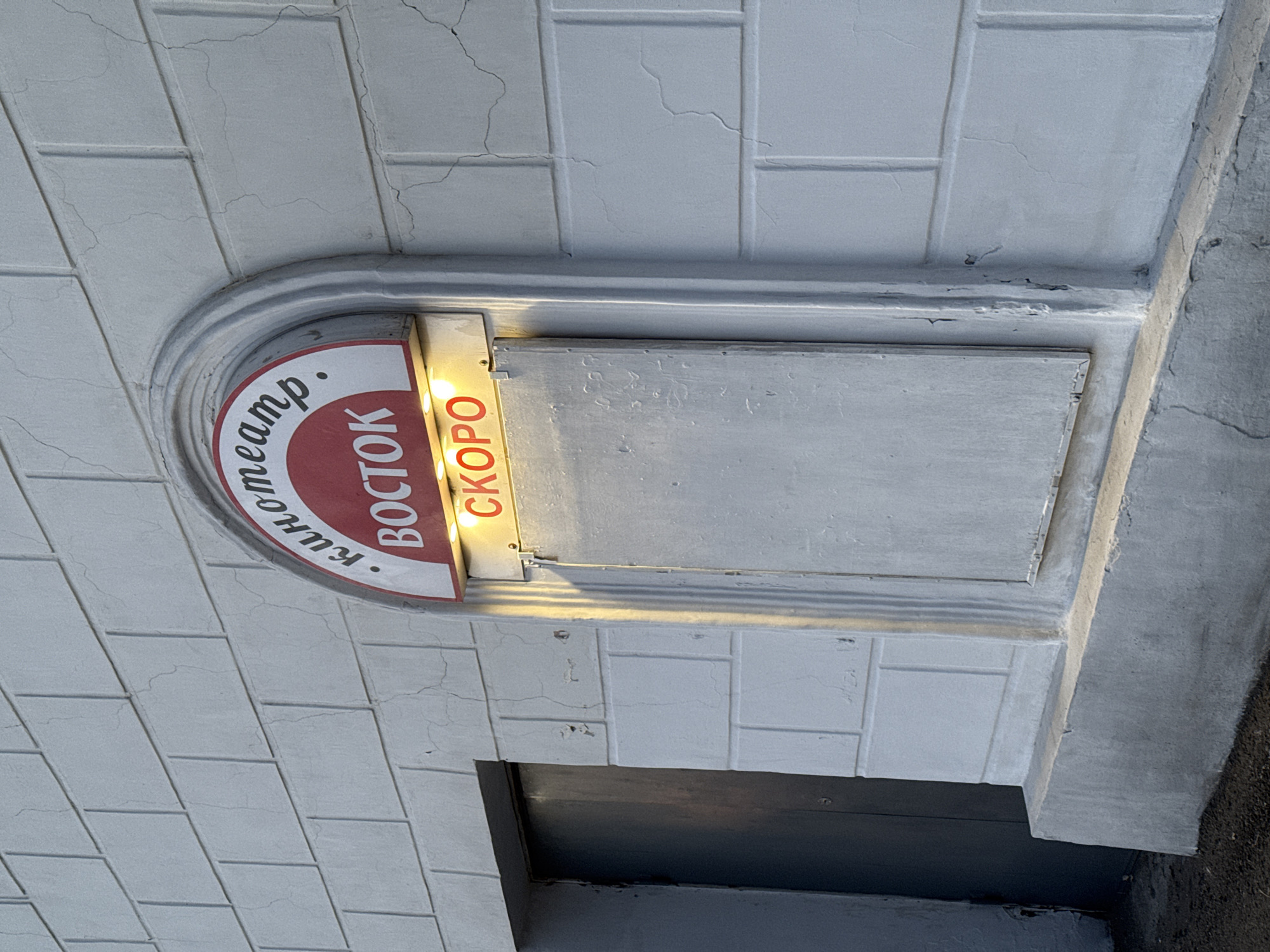 The image shows a sign for the "Vostok" (Восток) cinema in Borzya, Russia. The sign is attached to a gray, brick-patterned wall above a dark doorway or entrance. The sign is semi-circular with the words "Кинотеатр. Восток" (Kinoteatr. Vostok, meaning Cinema. Vostok) in white lettering on a red background, with "Кинотеатр" curving along the top and "Восток" written vertically. To the right of the curved sign is a rectangular section with the word "Скоро" (Skoro, meaning Soon) written vertically in red. The rectangular section also appears to have a cover that could be pulled or slid to cover the sign. The sign is lit from within, illuminating the word "Скоро". The overall appearance is somewhat aged and weathered. A concrete ledge or molding is present on the right side of the sign, extending down the side of the doorway. The lighting conditions suggest an outdoor setting with soft, diffused light.