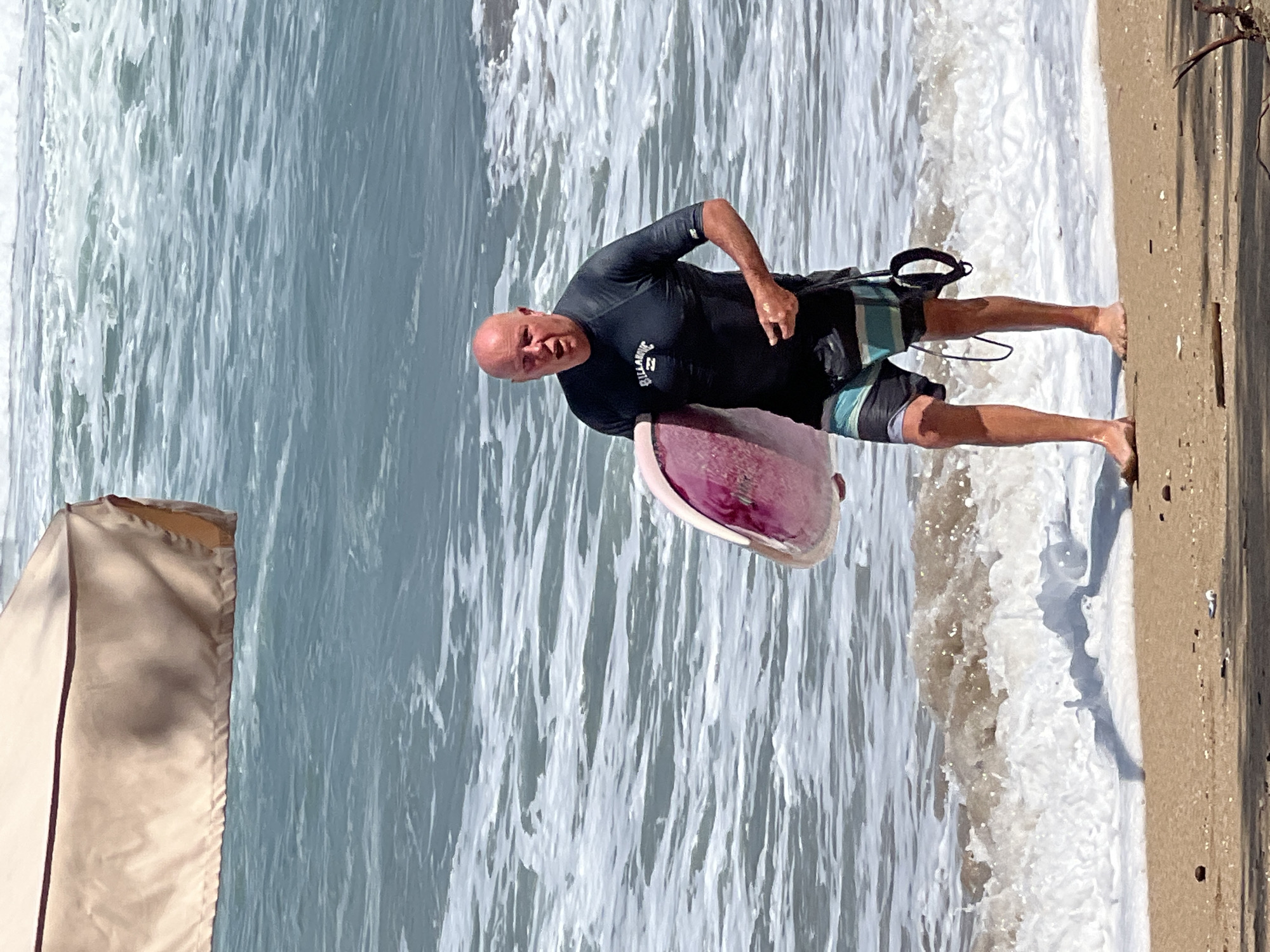 An older, bald Caucasian man wearing a black rash guard and striped swim trunks walks out of the ocean carrying a pink and white boogie board. He is on a sandy beach with waves and white foam washing ashore. In the upper left corner, a beige beach umbrella is visible. The scene appears to be Kuta, Indonesia.