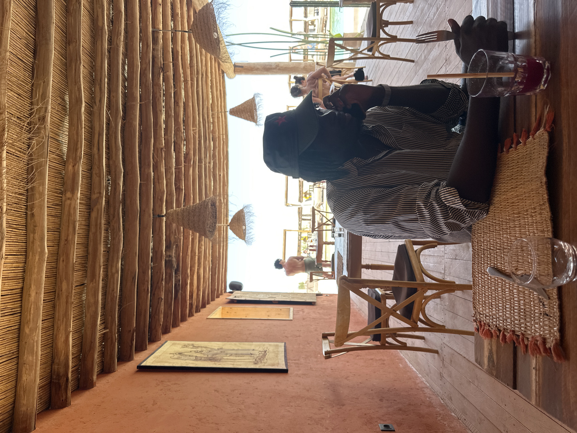 A person is seated at a table at a restaurant in Sidi Badhaj, Morocco. The person is wearing a striped shirt and a cap with a red star. They are holding a fork and a knife. A glass of red juice is on the table. A straw is in the glass. The restaurant has a thatched roof and orange walls. There are other people visible in the background. The table is set with woven placemats. A glass is empty on a place setting.