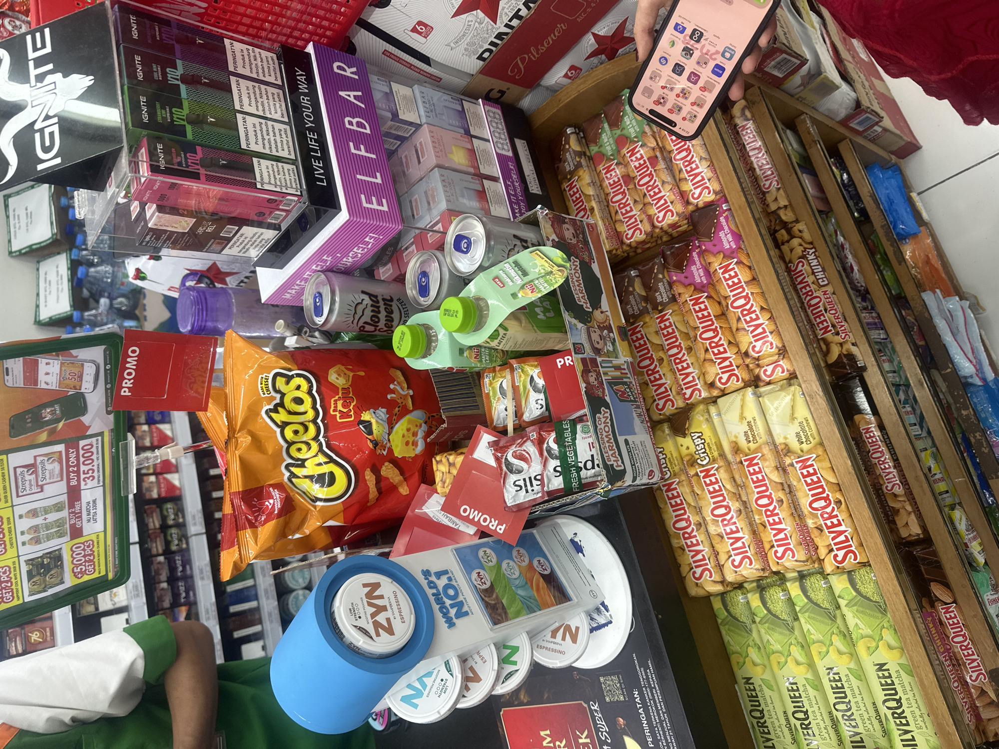 A person wearing a green shirt with orange and white details is partially visible in the lower-left corner. Next to them is a display of consumer goods in a retail setting. Dominating the scene are packages of Cheetos, Silver Queen chocolate bars in various flavors (green tea matcha, almonds, fruit & nuts, and chocolate with cashews), ZYN nicotine pouches in a blue container, and Elfbar and Ignite vaping products. Also present are cans of Cloud Seven seltzer, Nu Matcha Lattea drink, and small packages of seasonings.
A hand holding a smartphone with a variety of app icons on the screen is visible in the upper-right corner. There are promotion cards for several products on display, along with a pricing sign.
The store appears to be well-stocked, offering a wide array of snacks, beverages, and nicotine products. It could be located in North Kuta, Indonesia as indicated by a beer carton in the background.