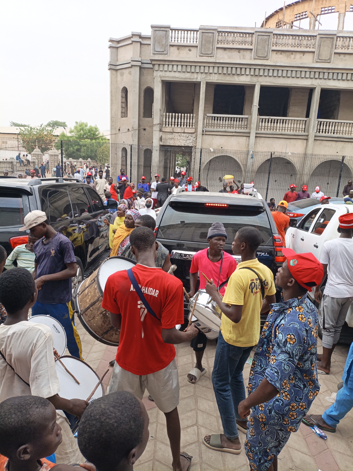 A daytime scene in Tudun Wada, Nigeria, shows a crowded street in front of a large, unfinished building with arched windows and a balustraded balcony. The street is filled with people, many wearing red hats and colorful clothing. A group of young men are playing drums, creating a festive atmosphere. Several black SUVs are parked nearby, and more people are visible in the background, some sitting on a low wall. The environment suggests an urban area, possibly a gathering or celebration.