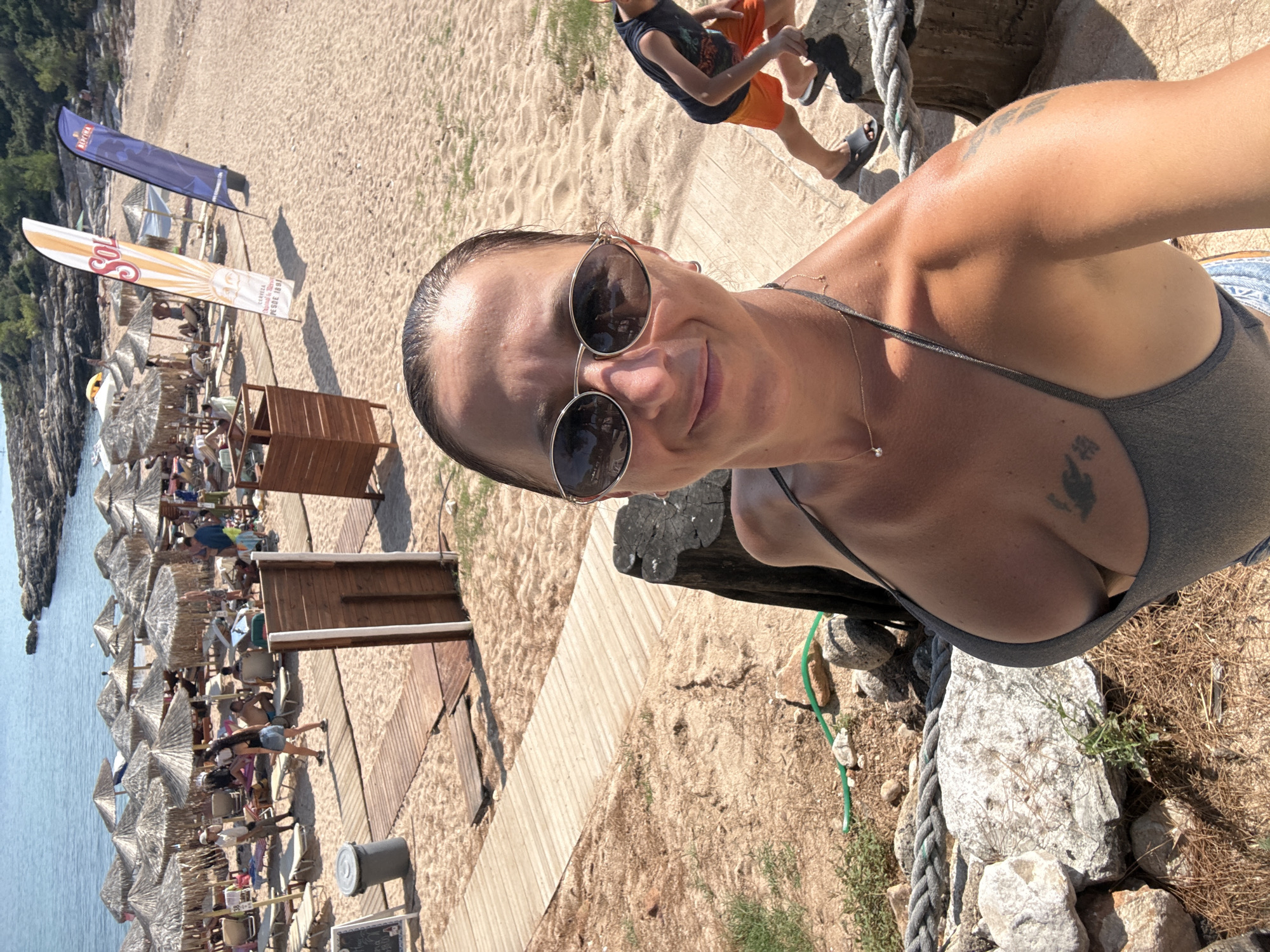 This is a first-person perspective, medium shot, outdoor photograph taken on a sunny day at Potos, Greece. The primary subject is a woman, who is posing for a selfie. She is wearing a gray bikini top and sunglasses. Her hair is wet and pulled back. In the background, a sandy beach is visible, lined with straw umbrellas and lounge chairs. Several people are seen relaxing on the beach. Wooden walkways are present, leading through the sand. A body of water, likely the sea, is visible on the left side of the frame. Two flags are planted in the sand, one blue with white text and another orange and yellow with text. The image conveys a relaxed, summery atmosphere. The time of day appears to be daytime, likely late morning or early afternoon, given the bright sunlight and strong shadows.