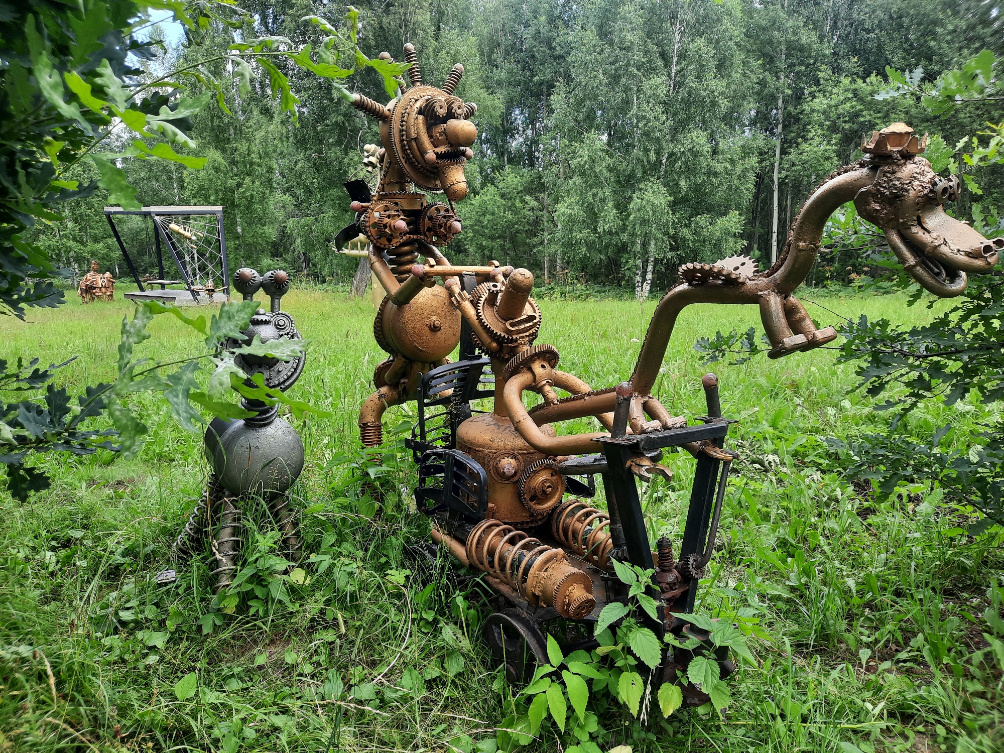 This image depicts a whimsical outdoor art installation made of scrap metal, located in the city of Zelenogorsk, Russia. The primary subjects are two distinct steampunk-inspired sculptures.
The larger sculpture in the center is a metal contraption resembling a person riding a vehicle. The "person" is constructed from various metal parts, including gears, pipes, and what appears to be a large, round metallic body. This figure is seated on a vehicle with wheels and is holding onto handlebars. The vehicle itself is also made of metal components, with visible springs and gears.
To the left of the main sculpture is a smaller, grey metallic figure. This figure is standing and has a more robotic appearance, with a round body and coiled metal legs.
To the right of the central sculpture, a long, serpentine metallic structure extends upwards, forming a head that resembles a dragon or dinosaur. This head is detailed with gears and other metallic pieces, and it is adorned with a crown-like object.
The sculptures are situated in a grassy outdoor environment, with lush green grass and various plants growing around them. In the background, a dense forest of trees can be seen, suggesting a natural or park-like setting. A metallic swing set structure is visible in the middle ground to the left. The overall scene is bright, with natural lighting. No people are present in the image.