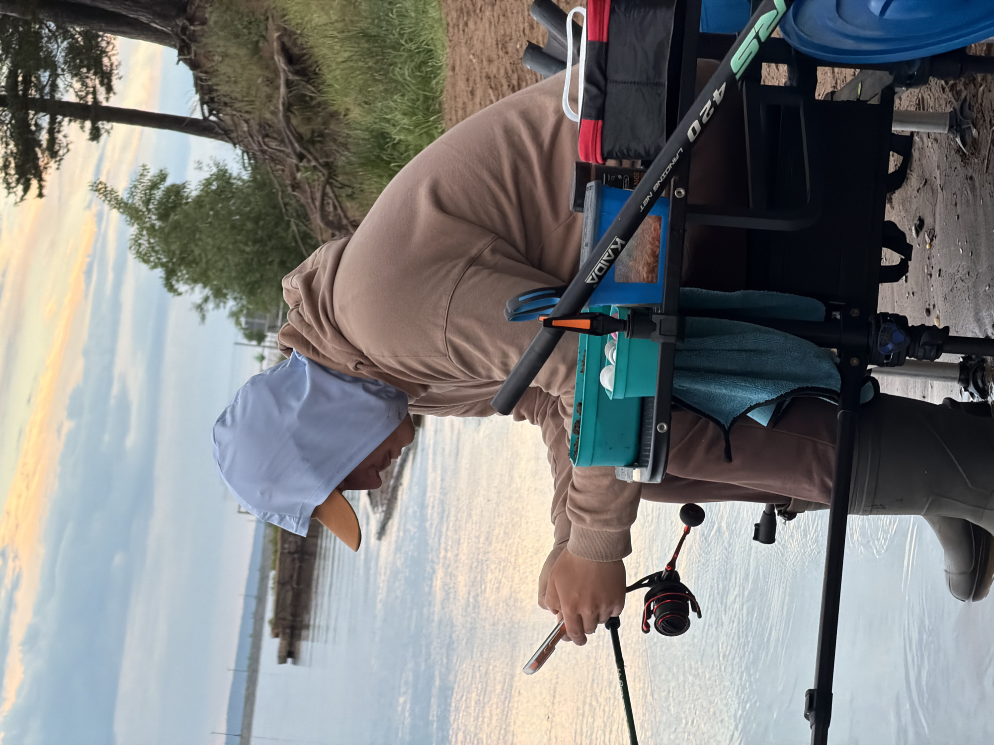 A person in a light brown hooded sweatshirt and a light blue hat is fishing by a body of water. The person is seated in a black fishing chair with various fishing accessories attached. The fishing rod is in their hands. A tree is visible on the left, and a pier is in the background across the water. The location is likely near Chaikovsky, Russia, based on available information about the user.