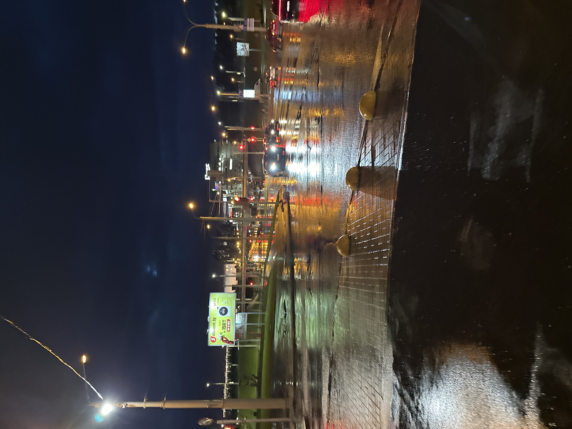 The image depicts a night scene in Minsk, Belarus, showcasing a wet street with reflections of city lights and car headlights. A billboard advertising "5 element.by" is visible on the left side of the frame. Streetlights illuminate the road and nearby structures. Cars are stopped at a traffic light further down the street. The pavement is wet, reflecting the light. Yellow bollards separate the road from the sidewalk. The sky is dark.