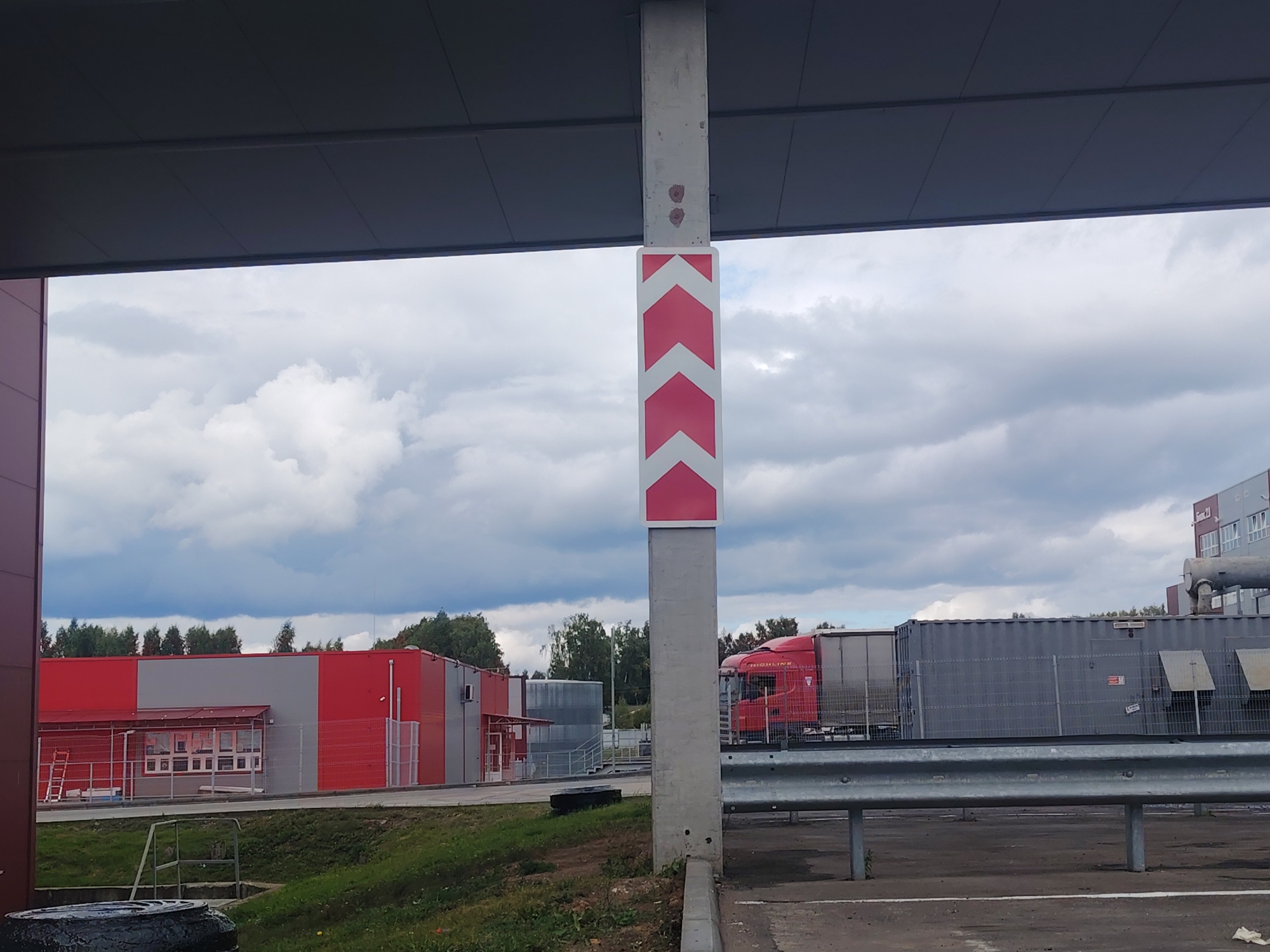 A concrete pillar with a chevron traffic marker is prominently featured in the foreground, partially obscuring a red and gray industrial building to the left and a gray storage container and a red truck to the right. The pillar, a light gray color, has three red and white chevron markers indicating a turn or a directional change. Above the chevron markers, there are two reddish-brown smudges on the concrete. The industrial building on the left is long and low with a gray roof over a section. A red ladder leans against the left side of this building. Behind the industrial building, a line of green trees is visible under a cloudy, overcast sky. To the right of the pillar, a large gray storage container is partially visible, and a red truck with "HIGHLINE" printed on its side is parked in front of it. A silver metal guardrail runs across the middle of the image. The ground in front of the guardrail is paved with asphalt, and the ground behind it consists of grass. The scene appears to be an outdoor industrial or logistics area.