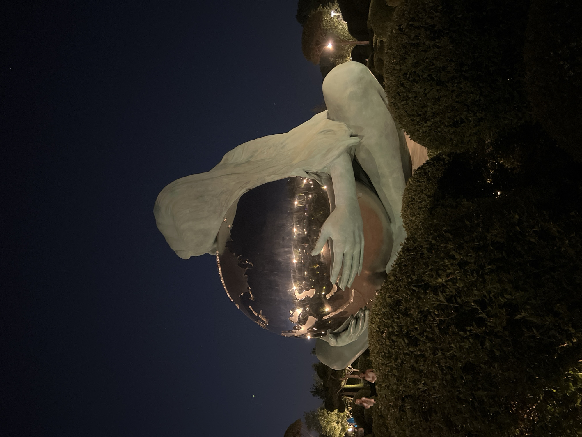 A large, bronze statue of a woman is depicted at night, holding a shiny, spherical object that appears to be a globe. The statue is seated with its legs crossed, and its long hair flows down its back and over its shoulders, partially obscuring the globe. The globe reflects the surrounding lights of what appears to be a park or garden. In the background, there are trees, bushes, and streetlights. Two individuals are visible in the middle ground, standing near the statue and looking towards it. One is wearing a light-colored top, and the other a white top. The sky above is dark blue and clear, with a few faint stars visible. The statue is positioned in an open area surrounded by manicured bushes. This statue, known as "The Child of the Earth," is located in the "Park of Culture and Recreation" in Krasnodar, Russia.