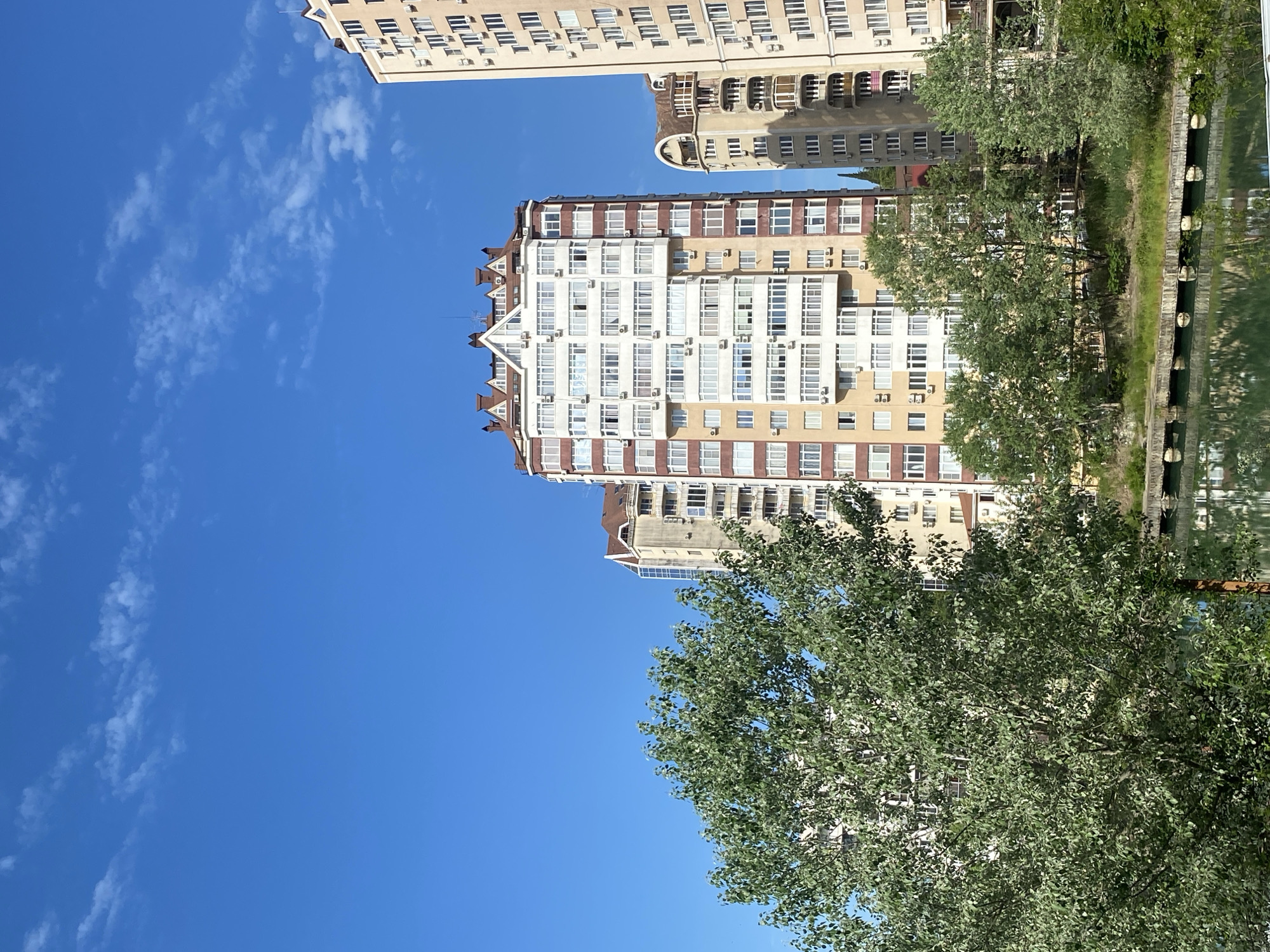 The image shows several apartment buildings in Sochi, Russia, viewed from a low angle, with trees partly obscuring them. The sky is blue with scattered white clouds. The buildings are light-colored with many windows and balconies. One of the buildings has a brown and white color scheme. There is a reflection of the buildings and trees in the water.