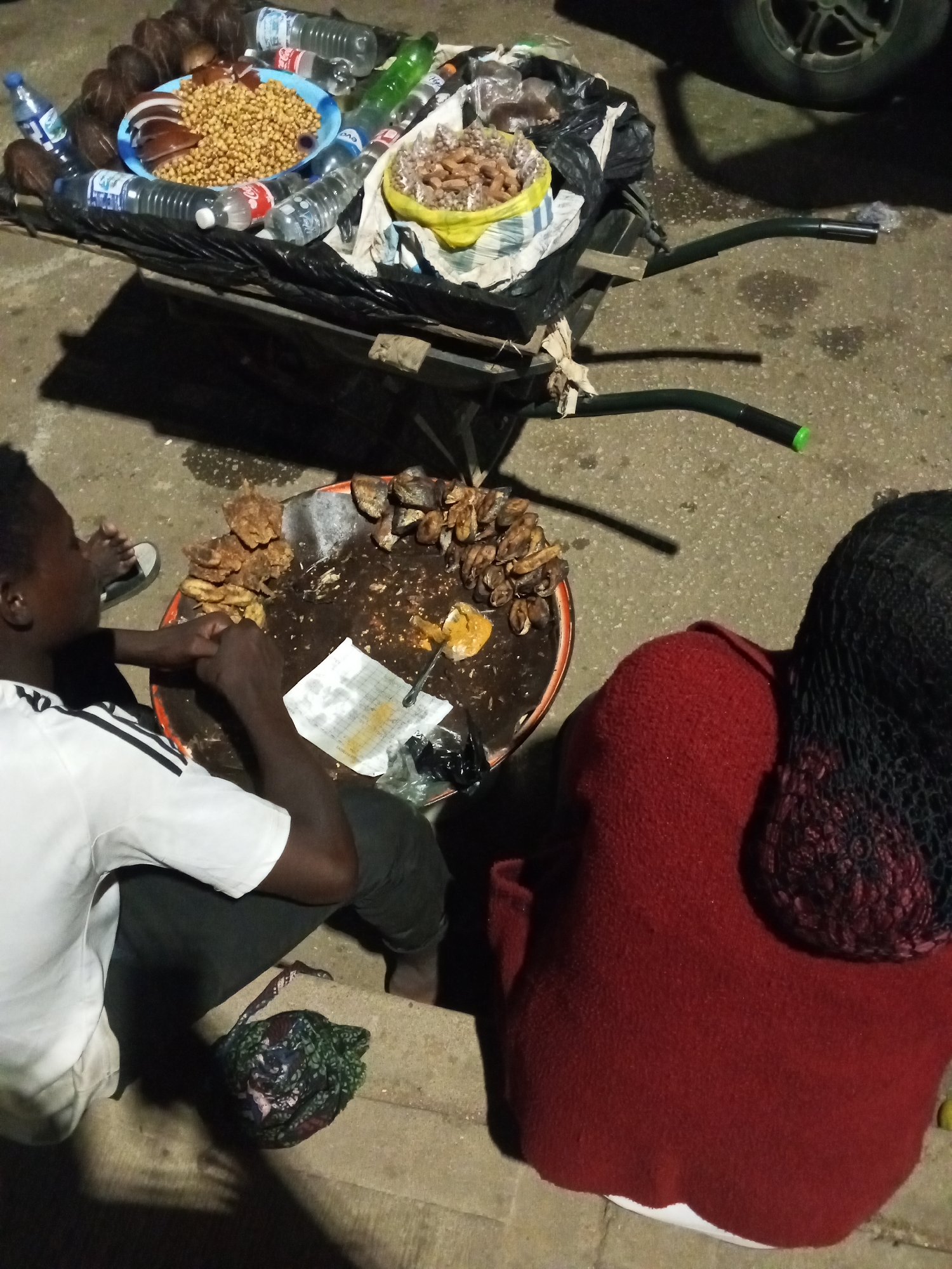 The image shows a street food vending scene in Bukuru, Nigeria. A wheelbarrow is loaded with coconuts, bottles of water and soda, and bowls of peanuts and other items. A man in a white t-shirt sits near a large tray filled with fried food items and a paper bag. Another person in a red sweater and a hair net is sitting on the side of the table. The scene takes place on a concrete surface, possibly a sidewalk, at night. A car tire is visible in the upper right corner.