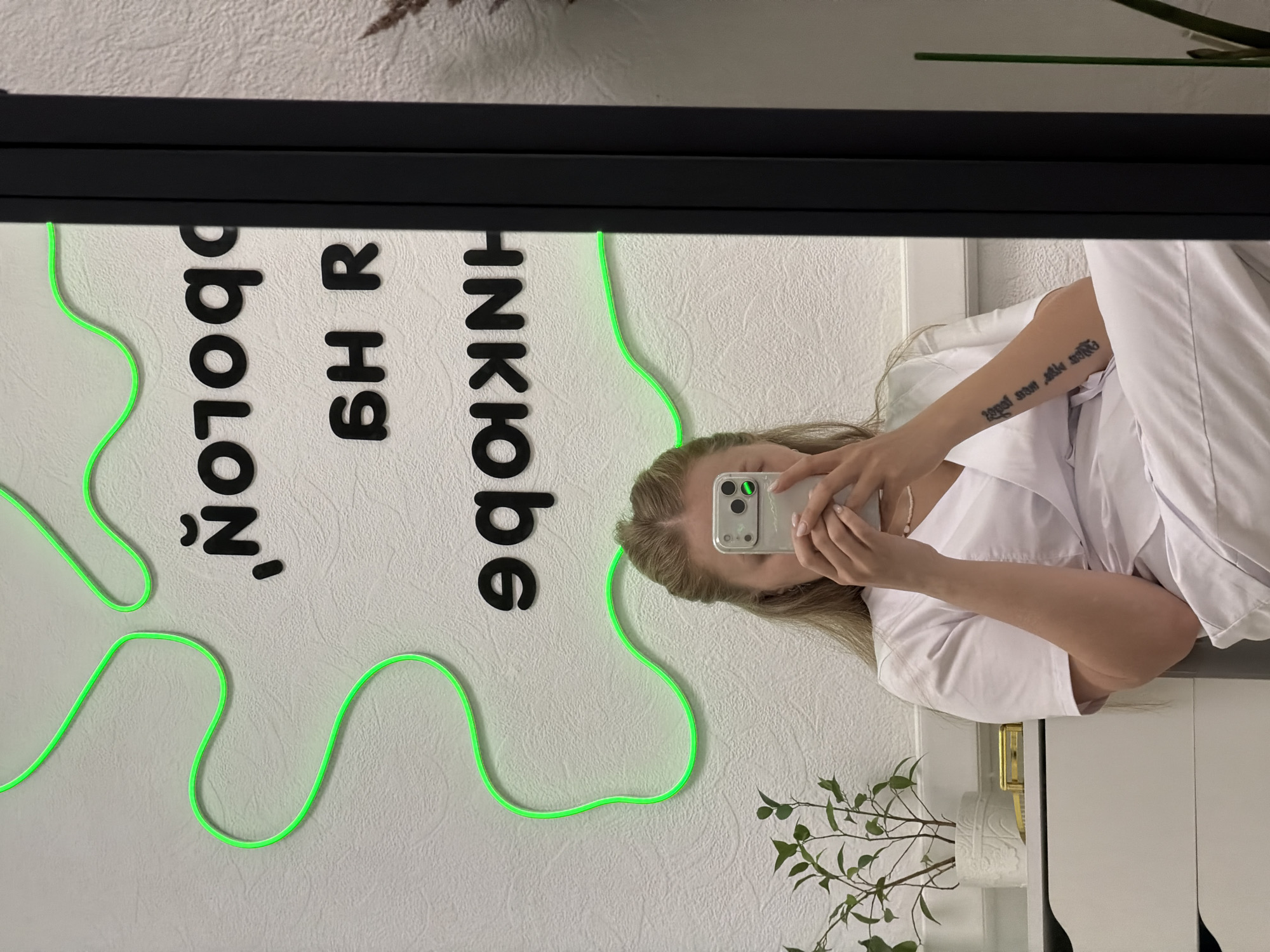 A photograph shows a mirror reflection. A woman in a white uniform is taking a picture of the reflection with her phone. The reflection shows a white wall with black lettering and green neon tubing forming a design, along with a plant. The lettering in the reflection is backwards and reads "HNKIobe, SHR, and 'NOLON'". The woman has blond hair, and her arm is visible in the reflection, with a tattoo on her forearm. The bottom portion of the mirror shows a white surface, possibly a cabinet or countertop, with a plant placed on it. Based on the lettering, the photograph was taken in Chaikovsky, Russia.