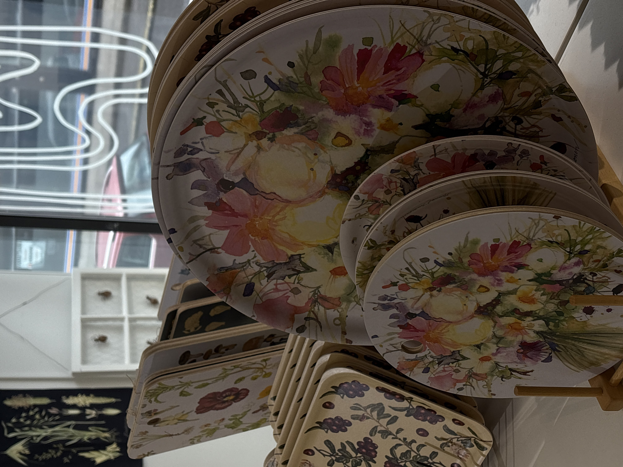 In the city of Pori, Finland, a retail establishment displays an array of decorative plates. The plates are stacked and arranged in a manner suggesting they are for sale. The primary focus is on the plates themselves, which are adorned with floral watercolor designs. One prominent plate in the foreground features a vibrant bouquet of pink, yellow, and white flowers with green foliage. Behind it, more plates are visible, showcasing different floral patterns and some with berry designs. To the left, a rack holds several rectangular trays with botanical illustrations. In the background, a white shelving unit with four compartments is visible, each containing a small object that appears to be a preserved insect. Further back, a window reveals an outdoor scene with a glimpse of a red car and the reflection of white neon signage. The overall setting indicates an indoor retail environment, likely a shop selling home goods or art.