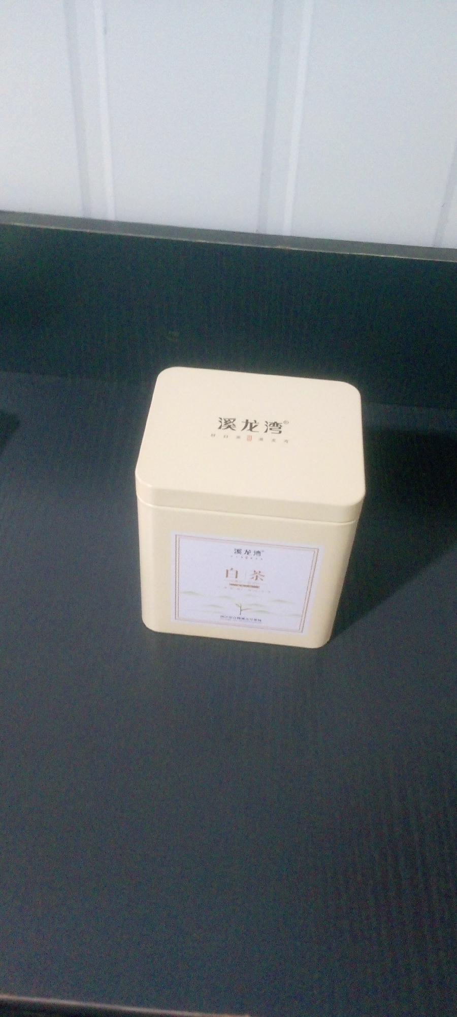 A square, cream-colored tin is centered on a dark, matte surface. The tin has rounded corners and a lid that appears to be slightly ajar. The lid features Chinese characters printed in black: "溪龙湾". Below these characters, in smaller print, is more Chinese text and the word "白茶" (White Tea). A decorative label with a faint landscape design and a white border is affixed to the front of the tin. The background consists of vertical, corrugated white panels and a dark horizontal surface. The lighting suggests an indoor setting. The city of Indo, Nigeria is not depicted in this image.
