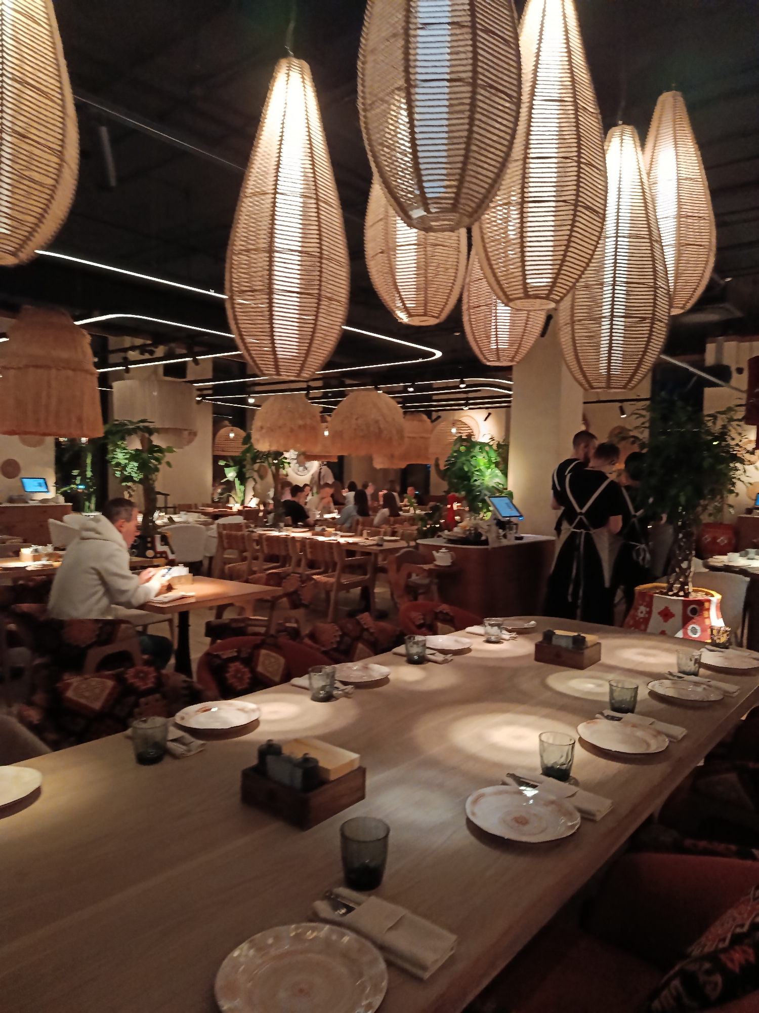The image shows the interior of a restaurant in Moscow, Russia. The foreground features a long, wooden table set with plates, silverware, glasses, and condiment holders. The seats are upholstered with patterned fabric. Above the table, numerous teardrop-shaped, woven pendant lights hang from the ceiling.
In the midground, other tables and chairs are visible, along with diners and restaurant staff. A man wearing a grey hooded sweatshirt is seated at one of the tables. Several employees dressed in black uniforms with white aprons are standing near a service area.
Green plants in decorative pots are placed throughout the restaurant. The overall ambiance appears to be dimly lit and upscale.