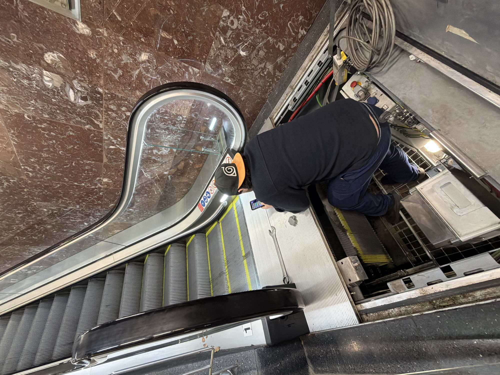 A man in a navy blue long-sleeved shirt and blue pants is kneeling and working on the inside of an escalator. The escalator is made of gray metal with yellow trim. The man is wearing a black baseball cap with an orange logo on the front. A wrench lies nearby on the metal surface. A phone is visible next to the man. The escalator is located in a building with a red marble wall in the background. Based on the architectural features visible, the escalator is likely located in Barcelona, Spain, although this cannot be confirmed with certainty.