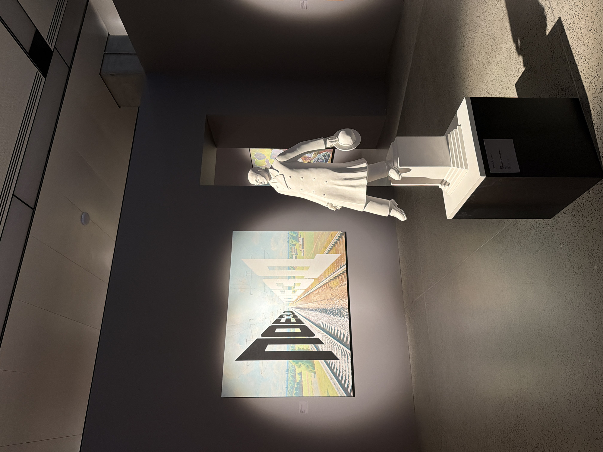 The image depicts an indoor exhibition space, likely a museum or gallery, with a minimalist aesthetic. In the foreground, a white statue of a man in a trench coat and hat stands on a dark pedestal. The statue is lit by a spotlight, casting a dramatic shadow on the floor. Behind the statue, a large, surreal painting is displayed on the wall. The painting features a railway track receding into the distance, with large, black and white three-dimensional letters forming words that appear to be embedded in the landscape. The walls are painted a deep gray, and the ceiling features modern lighting fixtures. The floor is a speckled gray concrete. The overall impression is one of a contemporary art exhibition. There are no visible people in the scene, and no specific Moscow landmarks are evident.
