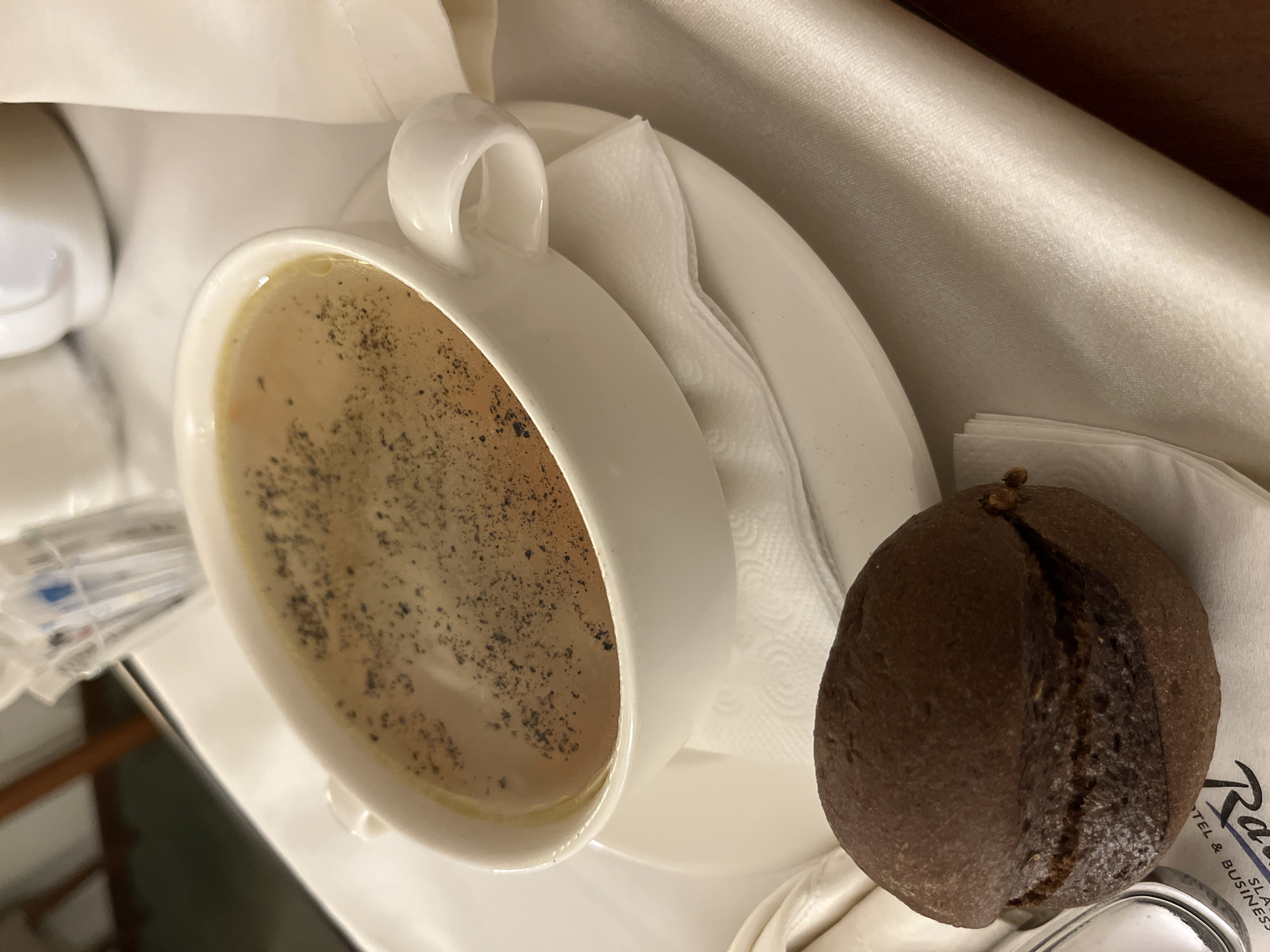 A high-angle shot depicts a cup of coffee served with a bread roll on a white surface. The coffee is in a white ceramic cup with a visible handle, placed on a white saucer. The coffee appears to have a light brown color with visible dark specks on the surface. Next to the cup, a dark brown bread roll rests on a white napkin. The napkin has a logo partially visible, indicating "Radisson" along with text relating to "Hotel & Business". There is also a packet of sugar next to the cup. The lighting is soft, casting subtle shadows. The background features a shiny white cloth. Given the Radisson logo, this image may be taken at a Radisson hotel, though the specific location in Moscow is not discernible from the image alone.