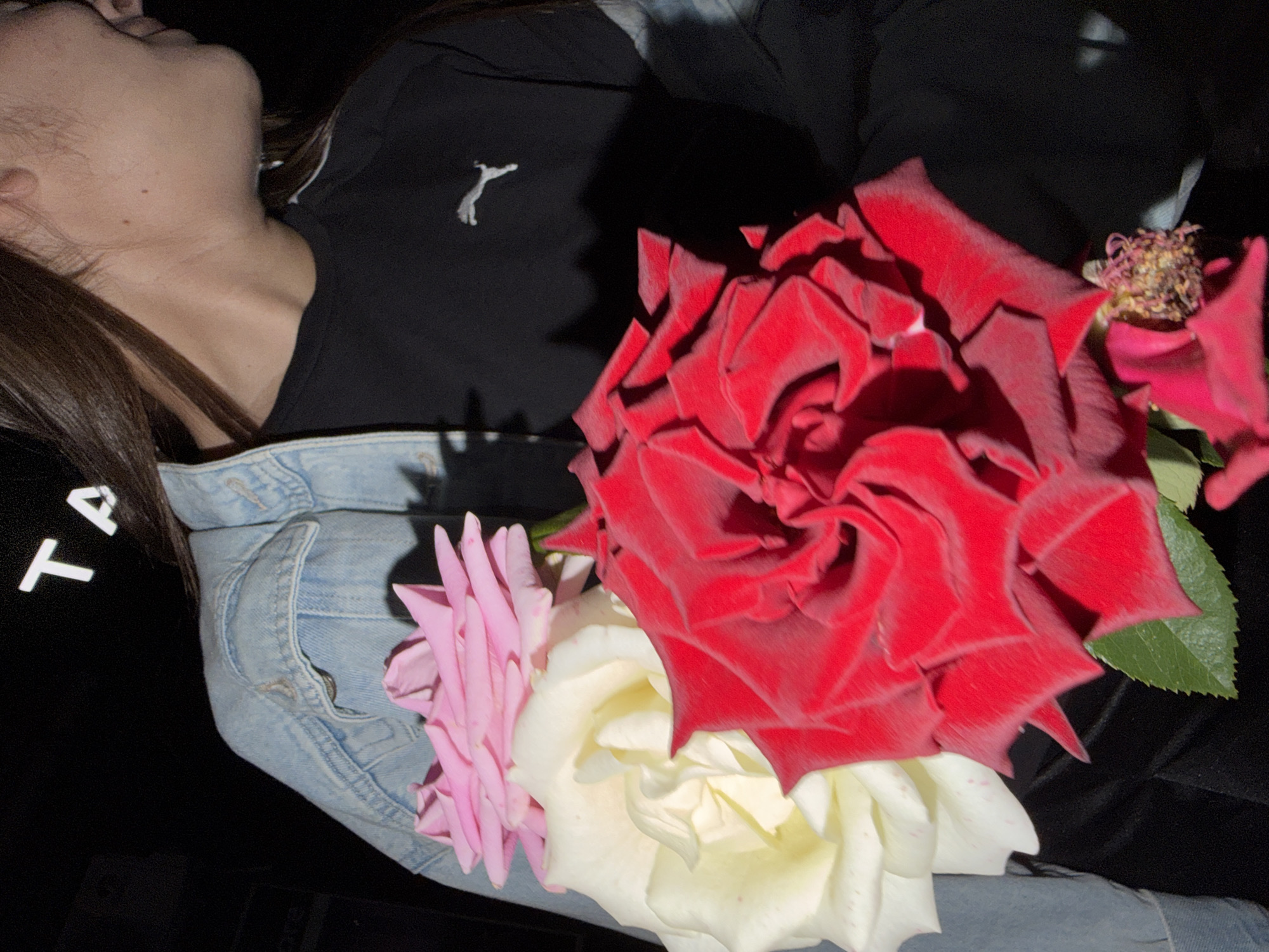 The image shows a close-up of a young woman holding a bouquet of three roses: one pink, one white, and one red. She is wearing a light blue denim jacket over a black t-shirt with a white Puma logo. The background is dark, with some text visible at the top which reads "TA". The scene appears to be indoors, possibly in a residential setting in Strășeni, Moldova. The lighting is artificial and creates strong shadows.