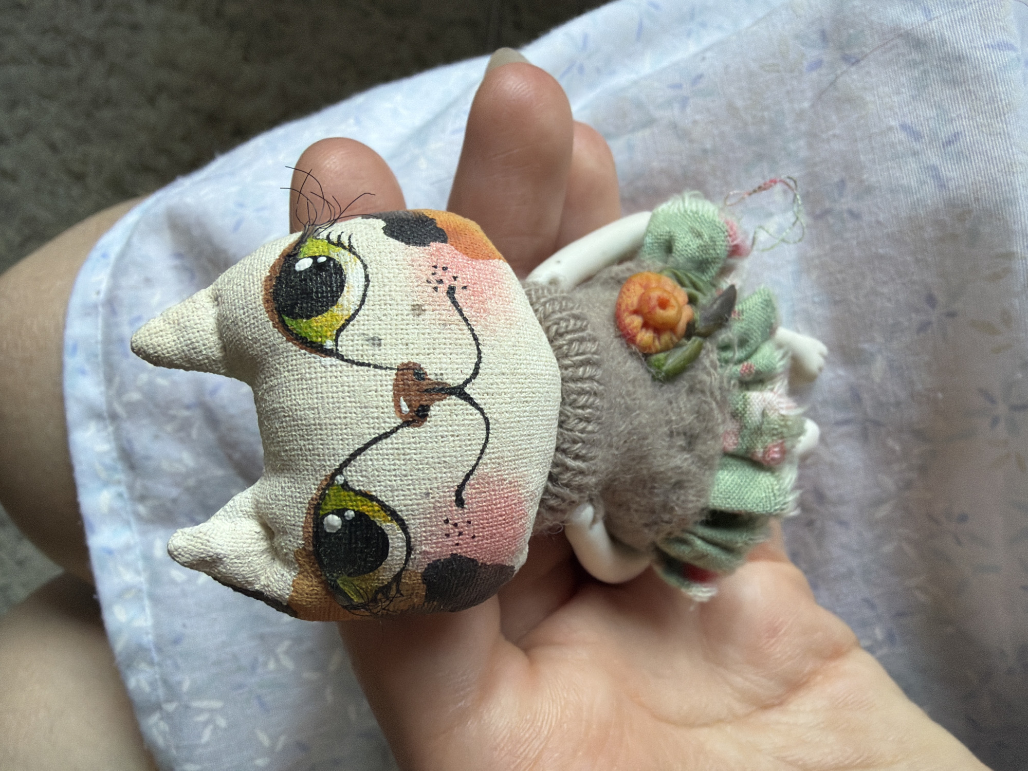 A person's hand is holding a small, handmade fabric doll that resembles a cat. The doll has large, green eyes with black pupils and yellow irises, painted eyelashes, a brown nose, and pink blush on its cheeks. It is wearing a grey knitted sweater over a green ruffled skirt decorated with small pink flowers. The doll is held in front of a light blue fabric with a subtle floral pattern, and a darker textured surface is visible in the background. There is no indication of the city of Chelyabinsk, Russia, in this image.