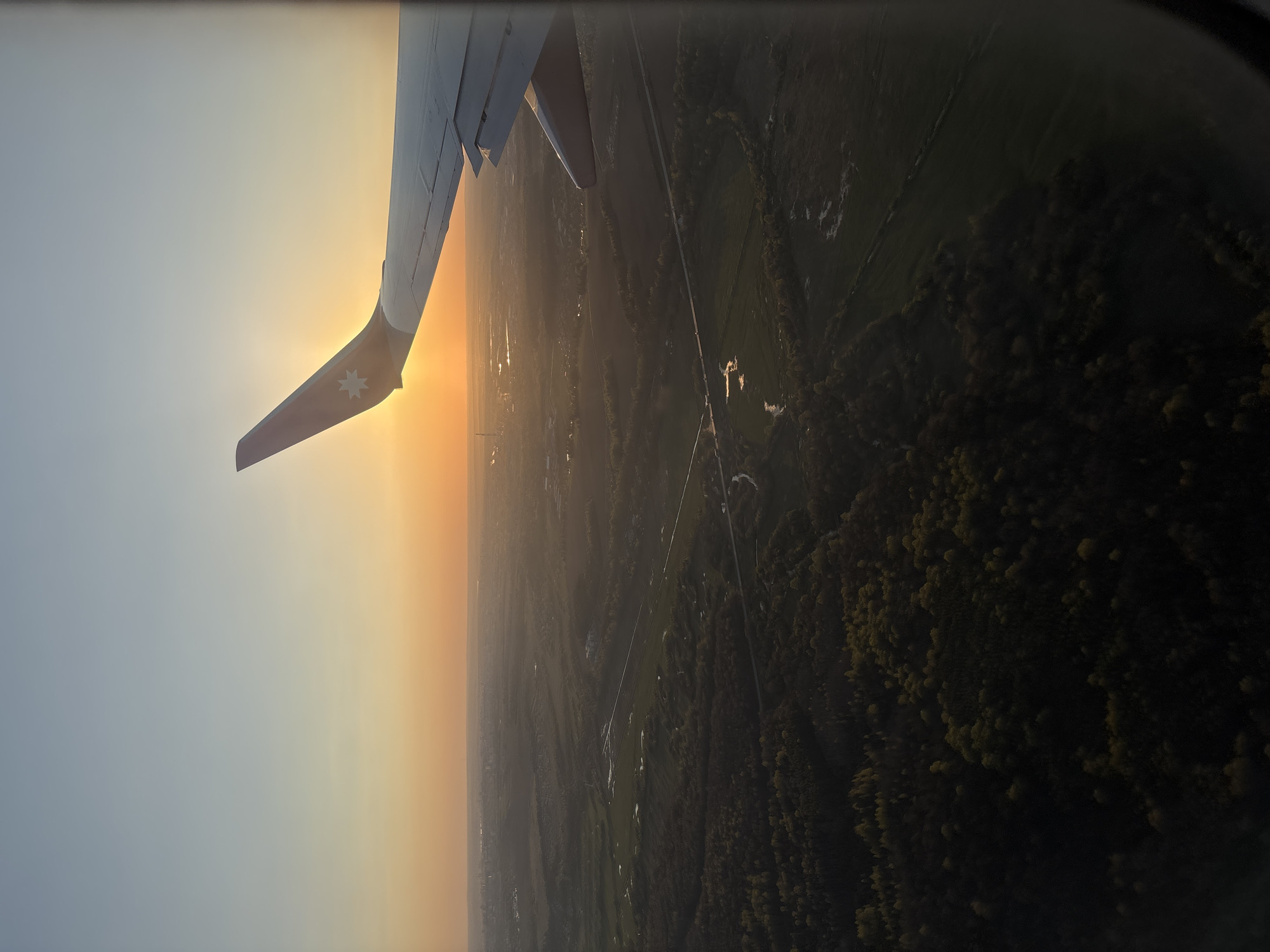 The image shows an aerial view from an airplane window, likely during sunset. A portion of the airplane's wing is visible in the foreground, bearing an Australian flag detail. Below, the landscape transitions from a hazy, yellowish-orange horizon to a green expanse of land. This terrain includes fields, patches of forest, and possibly small settlements. A straight road or utility line cuts across the landscape. There is no evidence to definitively determine this is Domodedovo, Russia. No people are visible.