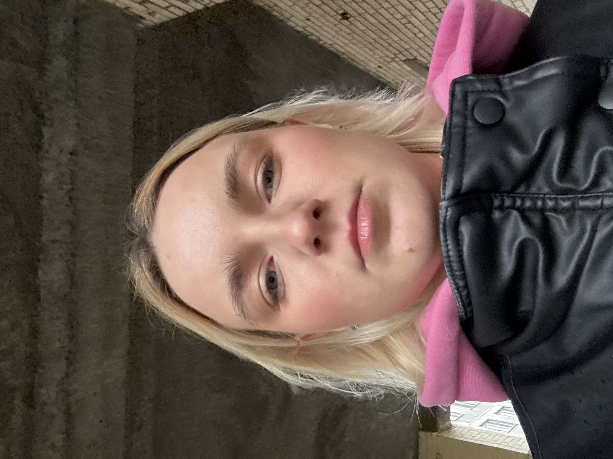 A close-up photograph depicts a woman in Unknown, Russia. She has light blonde hair, fair skin, and blue eyes. The woman is wearing a bright pink hooded sweatshirt and a black leather jacket. The background includes an aged brick wall, likely part of a building's exterior or interior. In the lower right corner of the image, the windows of another building are partially visible.