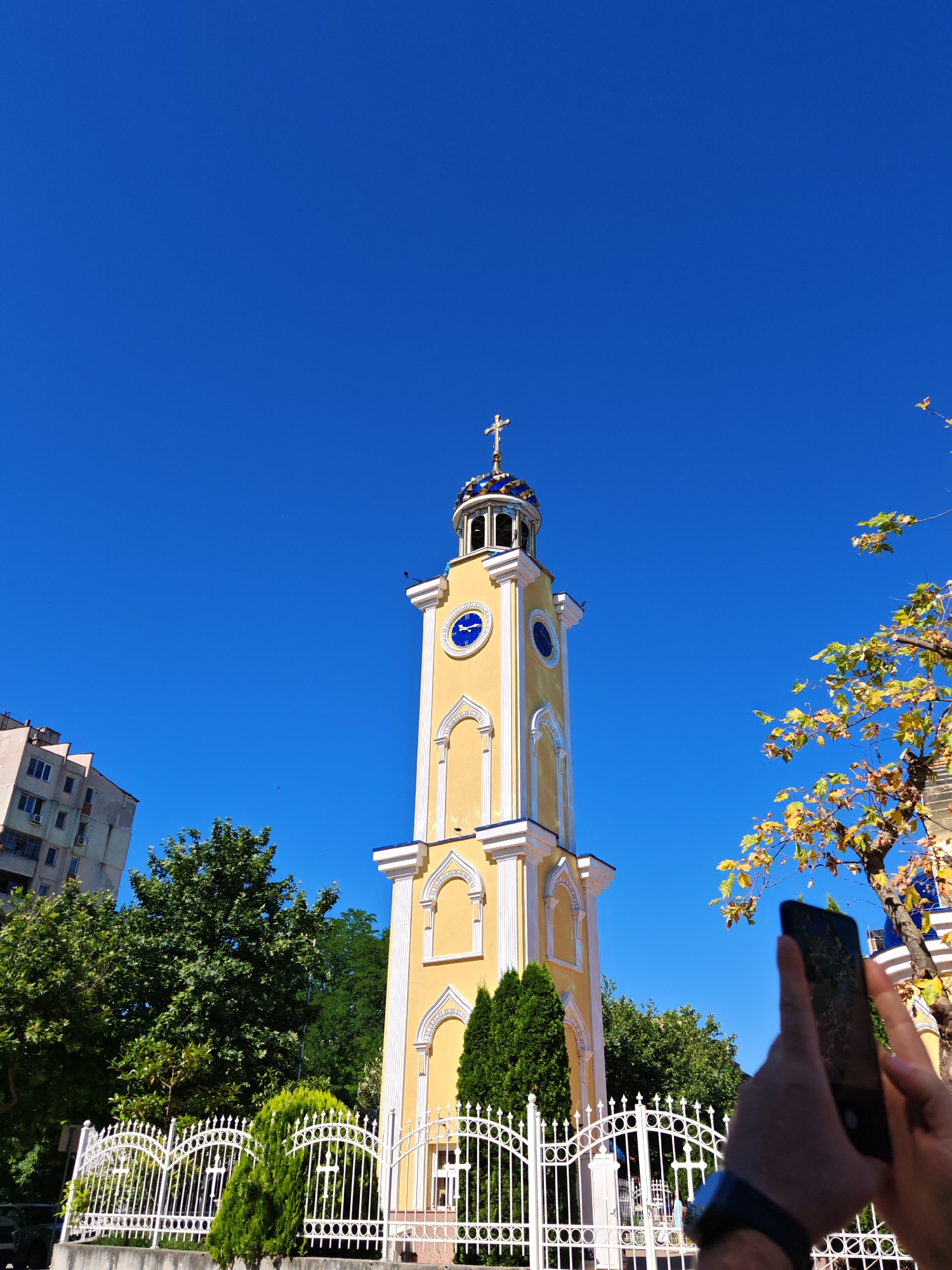 A low-angle, medium shot captures a yellow bell tower against a clear blue sky, with a person in the foreground holding a smartphone to take a photograph. The bell tower, a prominent architectural feature, is decorated with white trim and features a clock face with Roman numerals. A gold cross tops the tower, which is adorned with blue detailing. To the left of the tower, a tall, multi-story apartment building stands, and to the right, a tree with yellowing leaves partially obscures the view. In the lower portion of the image, a white, ornate metal fence runs across the scene, with trimmed evergreen bushes positioned behind it. The setting is likely within Plovdiv, Bulgaria, given the architectural style and urban environment. The scene appears to be captured on a bright, sunny day.