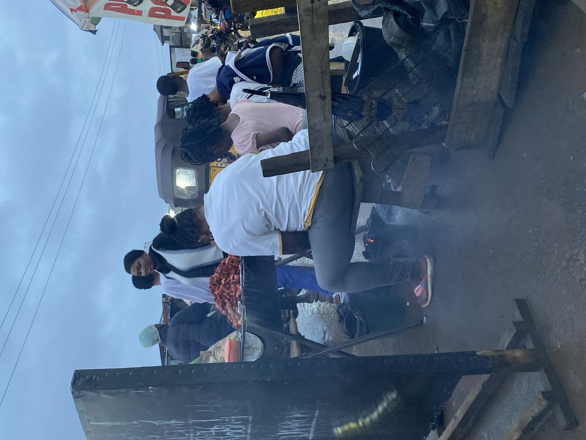 In Onibudo, Nigeria, a street scene unfolds under a cloudy sky. A charcoal grill, laden with what appears to be raw, marinated meat, sits on a makeshift stand, emitting a faint haze. Several individuals are gathered around, some attending to the food, while others stand nearby, observing. A woman in a white t-shirt and grey pants sits on a stool, facing away from the camera, seemingly engaged with the grill. Adjacent to her, a young girl in a pink shirt and a woman in a dark jacket with a white trim are also present. In the background, a yellow auto-rickshaw, a common mode of transport in the region, is visible, along with other market stalls and motorcycles, suggesting a bustling commercial area. The ground is wet, hinting at recent rain. A large, dark board with chalk markings stands to the left, adding to the informal market atmosphere.