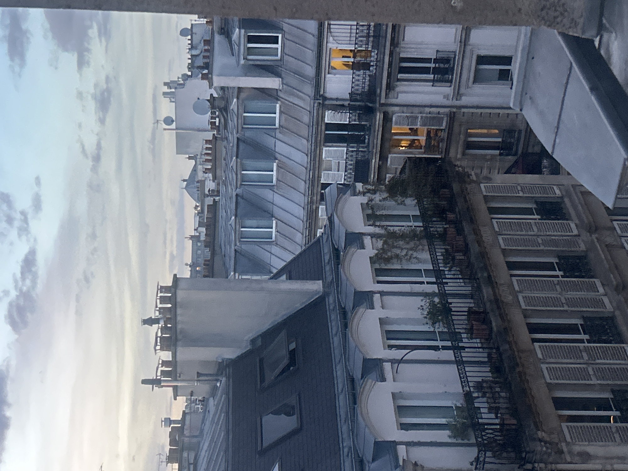 The image captures a vertical perspective of urban buildings, likely in an European city like Paris, under an overcast sky at dusk or early evening. The scene is dominated by multi-story residential or commercial structures with distinct architectural features.
In the foreground and middle ground, several buildings are visible, characterized by their light-colored stone facades, tall windows, and intricate wrought-iron balconies, some adorned with potted plants. Many windows have traditional louvered shutters, some closed, others open. Lights are visible in some windows, indicating interior illumination, which, combined with the soft, diffused light in the sky, suggests the approach of night.
The rooftops feature the iconic gray zinc material, typical of Parisian architecture, slanting steeply upwards. Numerous chimneys of varying designs pierce the rooftops, alongside modern additions like satellite dishes and air conditioning units. The sky above is a pale blue-grey with streaks of clouds, hinting at a quiet, calm atmosphere. No people or specific activities are discernible, creating a tranquil, architectural snapshot of an urban environment. There is no visible text in the image.