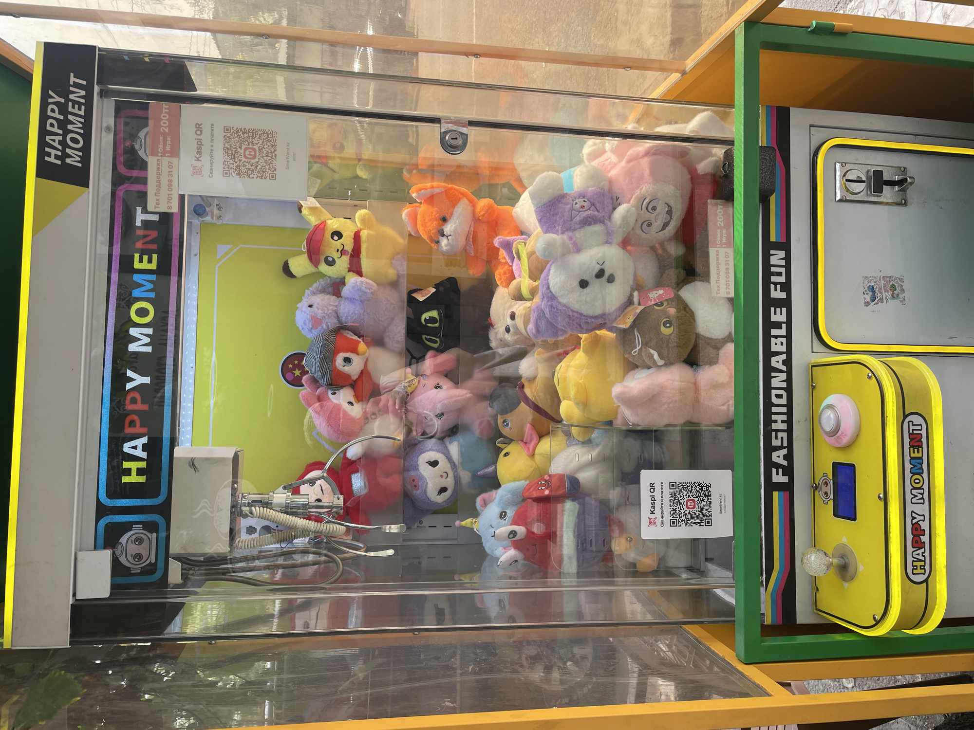 A bright yellow and green claw machine arcade game, labeled "HAPPY MOMENT," is positioned outdoors, likely on a street or in a park, given the dappled sunlight and foliage visible in the background. The machine is filled with an assortment of colorful plush toys, including Pikachu, various cartoon characters, and stuffed animals of different sizes and colors. A QR code for "Kaspi QR" is prominently displayed on the glass front, indicating payment can be made via this method. The control panel of the machine is visible at the bottom, featuring a joystick, a digital display, and a button. A coin slot is also visible on the lower part of the machine. The overall impression is one of a typical urban entertainment fixture, common in public spaces in cities like Almaty, Kazakhstan.