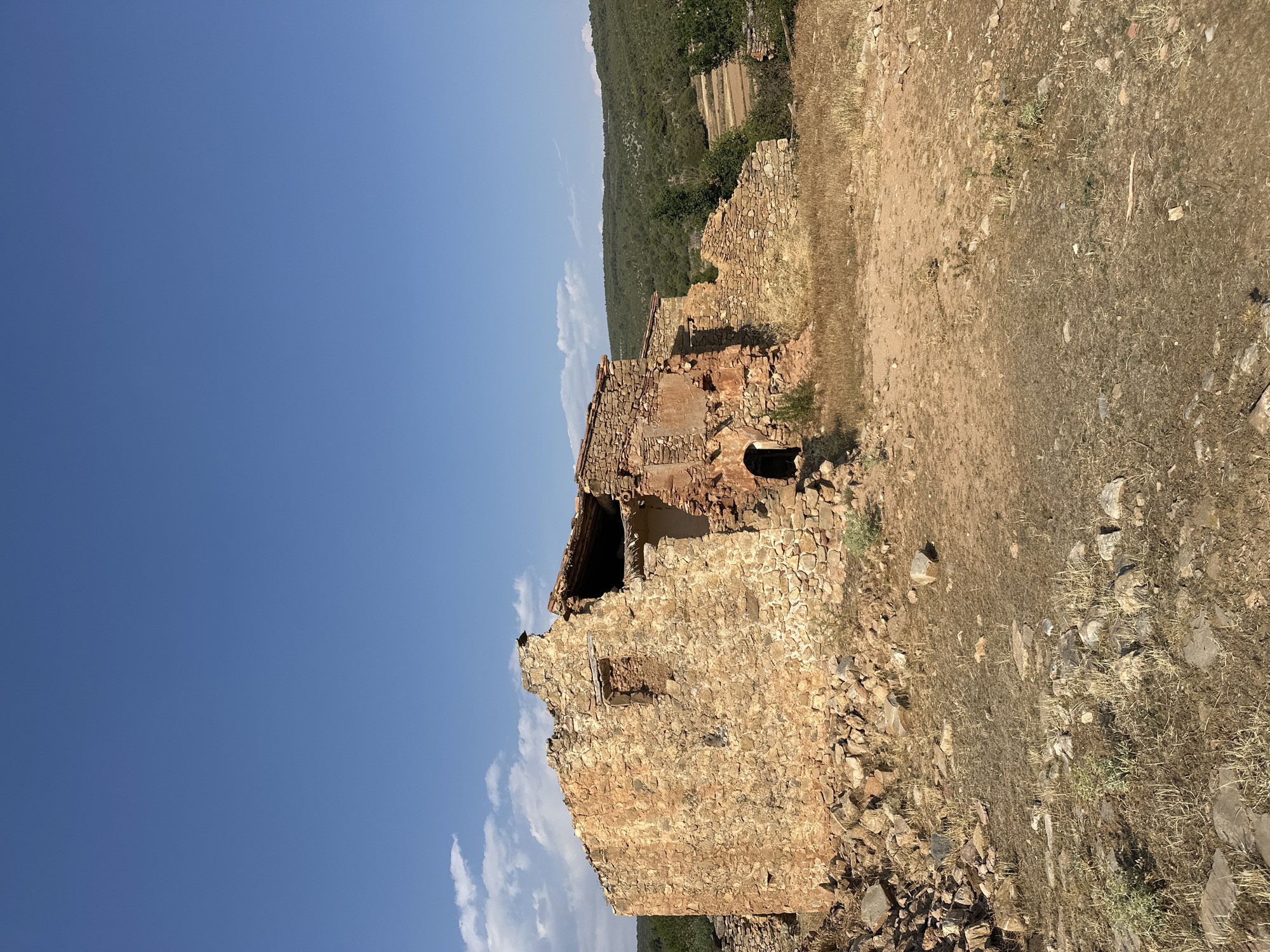 This is a vertical, outdoor, eye-level, medium shot of a ruined stone building under a clear blue sky with some scattered white clouds. The building appears to be old, with a partially collapsed roof and weathered stone walls. An arched doorway is visible on the side of the building. Dry, light brown grass and dirt cover the ground around the ruins. In the background, a lush green hillside rises, with trees and some cultivated terraces visible. The sun is shining, casting shadows, suggesting it is daytime, possibly late afternoon given the angle of the shadows. The scene evokes a sense of history and abandonment. There is no visible text in the image. The location context is Alpuente, Spain.