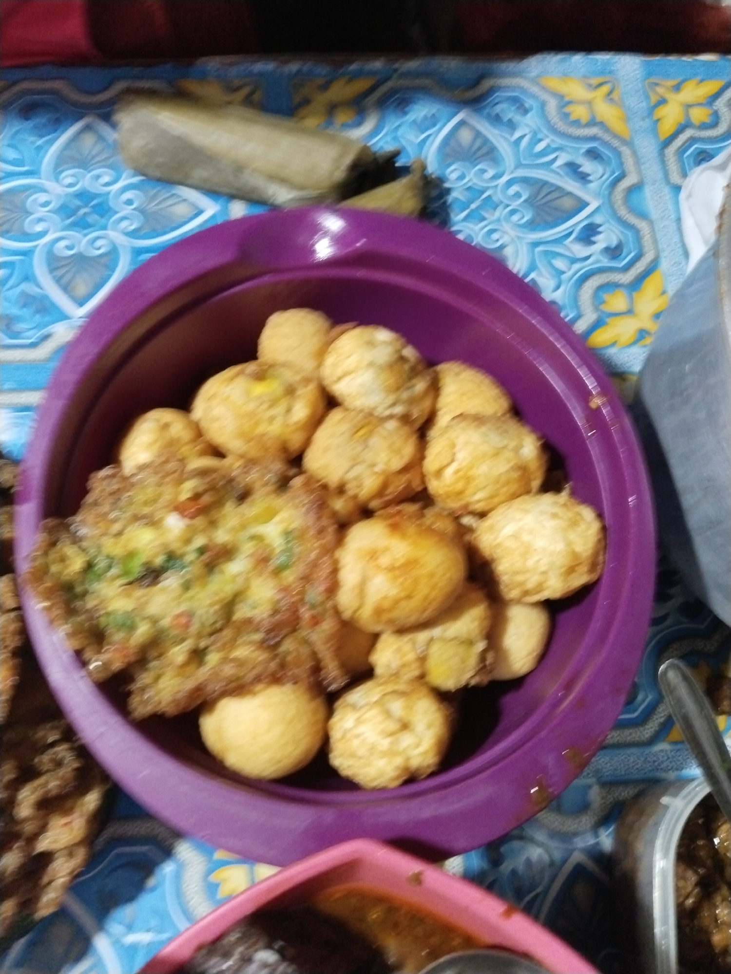 A close-up, overhead shot shows a purple bowl filled with fried food items. The bowl is on a blue and white patterned tablecloth. In the bowl, there are several round, golden-brown fried balls and one flat, golden-brown fried omelet-like item with visible green specks. To the right of the bowl, a clear plastic container holds a dark, saucy food item, and a metal spoon rests inside it. Part of a pink bowl is visible at the bottom of the image, also containing a dark food item. The background is out of focus but suggests a home environment.