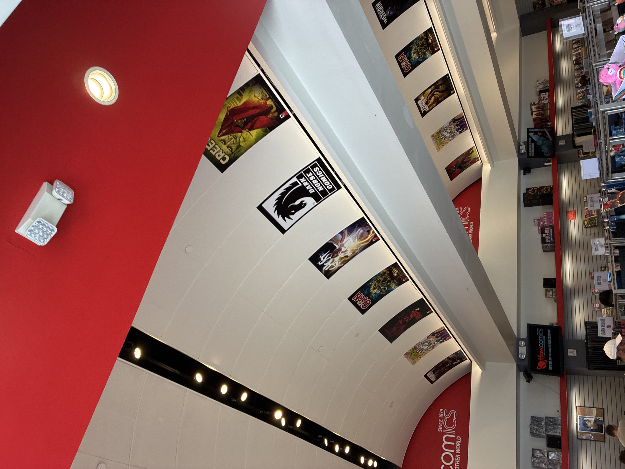 The image shows the interior of a comic book store. The ceiling is red and white with a curved design. There are lights along a black strip on the ceiling. Several comic book covers are displayed as banners hanging from the ceiling. On the wall below, a sign reads "tfaw.com" with a red logo and the text "WE'VE GOT JUST THE THING FOR YOUR COLLECTION." Below this, shelves are stocked with merchandise and more comic books. People are visible in the lower part of the image, browsing the merchandise. The store appears to be well-lit and organized. The location is identified as Unknown, United States.