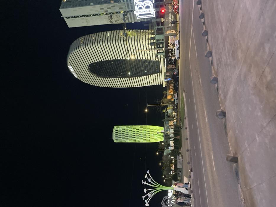 At night, a cityscape is viewed from ground level. Prominent buildings include a tall, elongated structure with a hollow oval shape, and a cylindrical building illuminated in green. Sidewalks border the street. Street lights illuminate the scene, and people are gathered near a large white floral sculpture. The buildings have text and logos, including what appears to be "Courtyard," and "Billionaire" displayed on their facades.