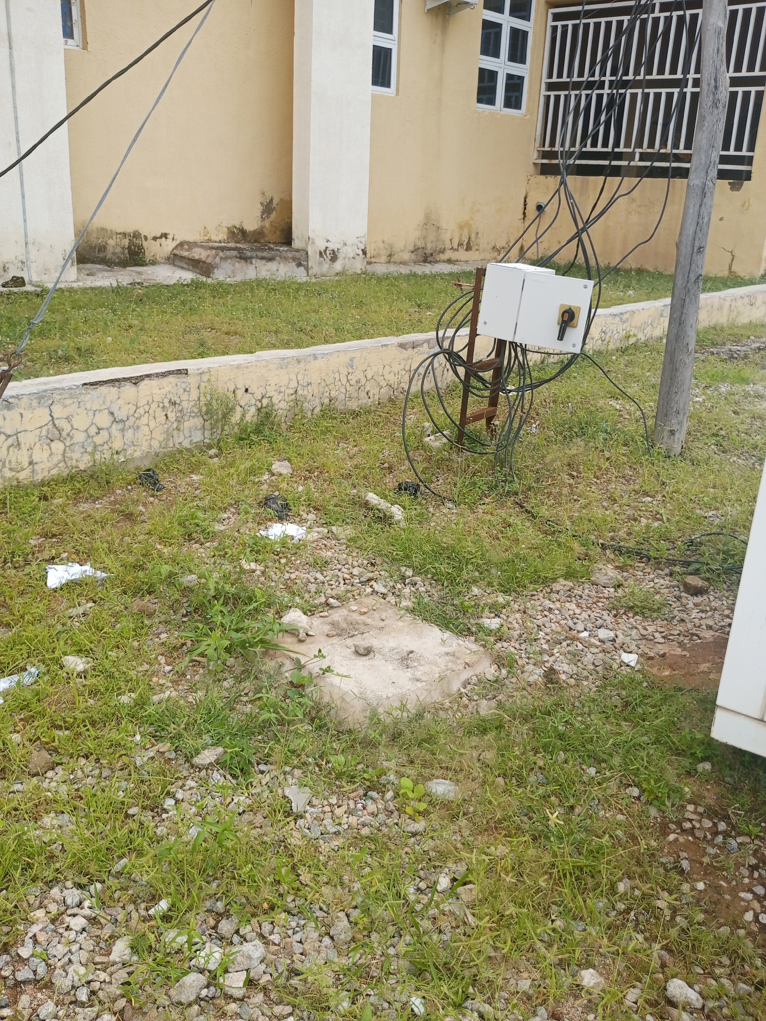 The image shows an outdoor scene in Jalingo, Nigeria, with a focus on electrical infrastructure and its surroundings. A light-colored building with cream-colored pillars and a beige facade is visible in the background. The building has several windows, some of which are barred with white metal.
In the foreground, a wooden utility pole stands prominently, supporting a white electrical box with a black switch. A tangle of black electrical wires is connected to the box and extends upwards and outwards, creating a messy but functional appearance. The box is mounted on a rusty metal frame that is anchored to the ground.
The ground is covered with a mix of green grass, gravel, and some scattered debris, including what appears to be white plastic bags and possibly some black plastic trash bags. A low, cracked concrete wall runs along the edge of the grass, separating it from the area where the building is situated. A concrete slab is embedded in the ground near the utility pole, possibly serving as a base for the pole or related equipment.
The scene appears to be during daylight, with overcast or diffused lighting, as there are no strong shadows. The overall impression is one of an urban or semi-urban environment with functional but somewhat unkempt electrical infrastructure. No people are visible in the image. There is no visible text.