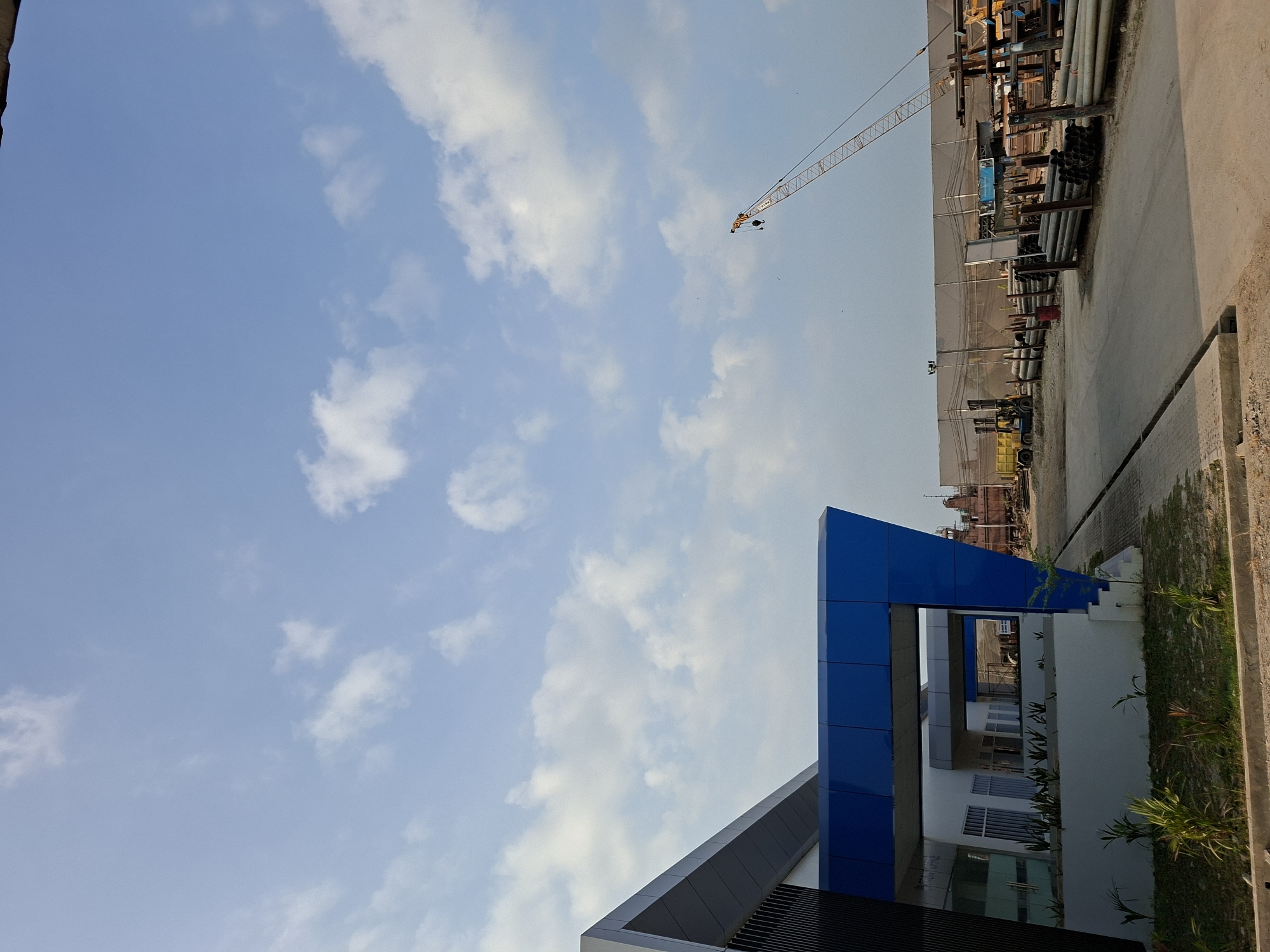 A low-angle shot captures a modern blue and gray building with a prominent blue overhang on the left side of the frame. The building appears to be an office or administrative facility. Beyond the building, a construction or industrial yard is visible. A tall, orange construction crane extends into the bright blue sky filled with scattered white clouds. The yard contains stacks of industrial materials, including pipes and metal structures, and a yellow forklift is parked in the distance. The ground is a mix of paved surfaces and dirt. The scene suggests an ongoing construction or industrial operation under a clear sky. The location is identified as Loa Buah, Indonesia.