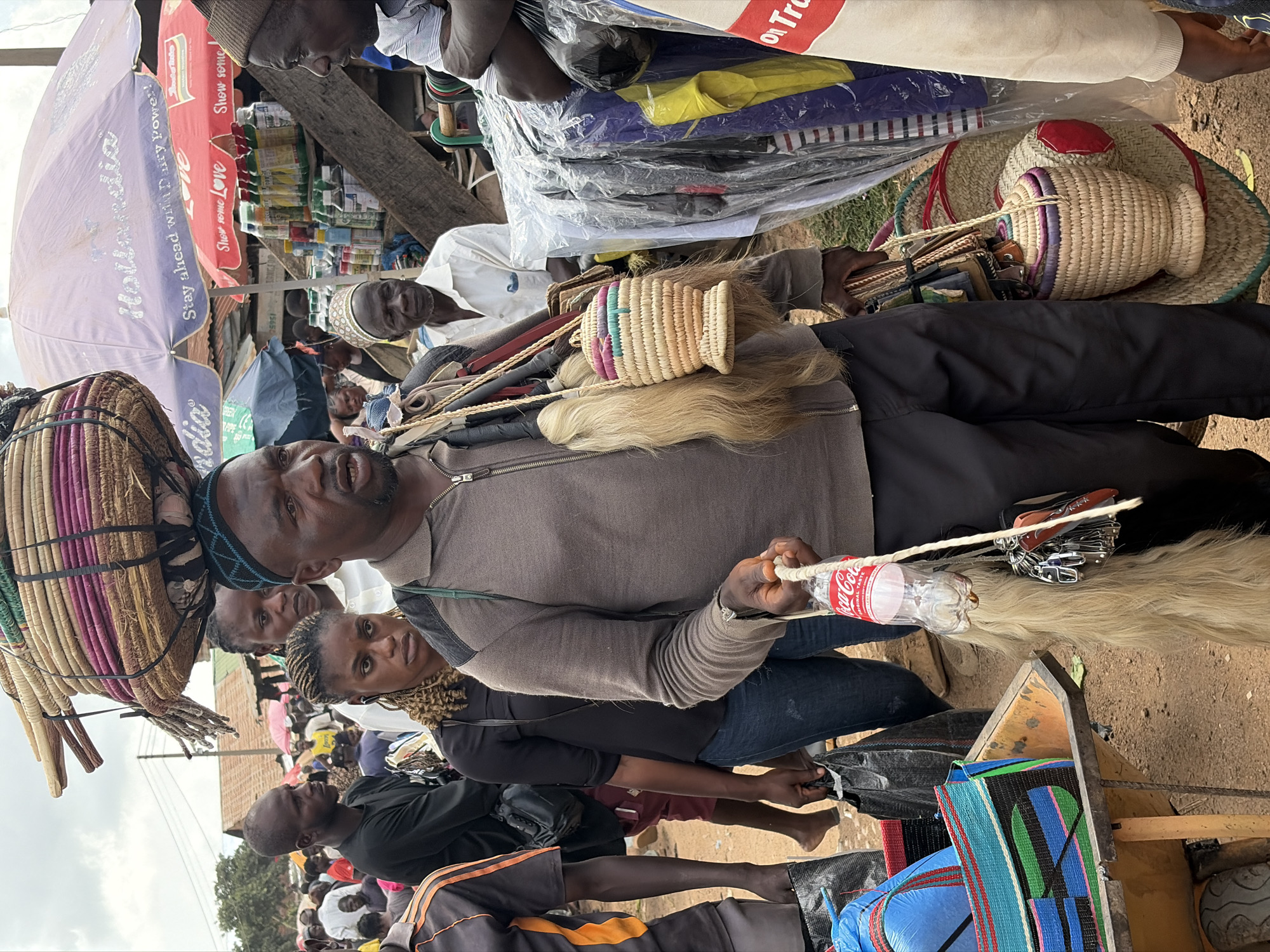 In Gura Top, Nigeria, a man with dark skin, wearing a gray zippered jacket and dark pants, carries a large woven basket stacked with items on his head. He is in a marketplace with other people and stalls. He holds a Coca-Cola bottle and a collection of keys and other items hanging from a rope in his right hand. A small woven basket hangs from his left shoulder. In the background, there are stalls with merchandise, including umbrellas advertising "Hollandia" and "Show some Love." The ground is dusty, and there are other individuals in the scene, some carrying bags and wearing hats.