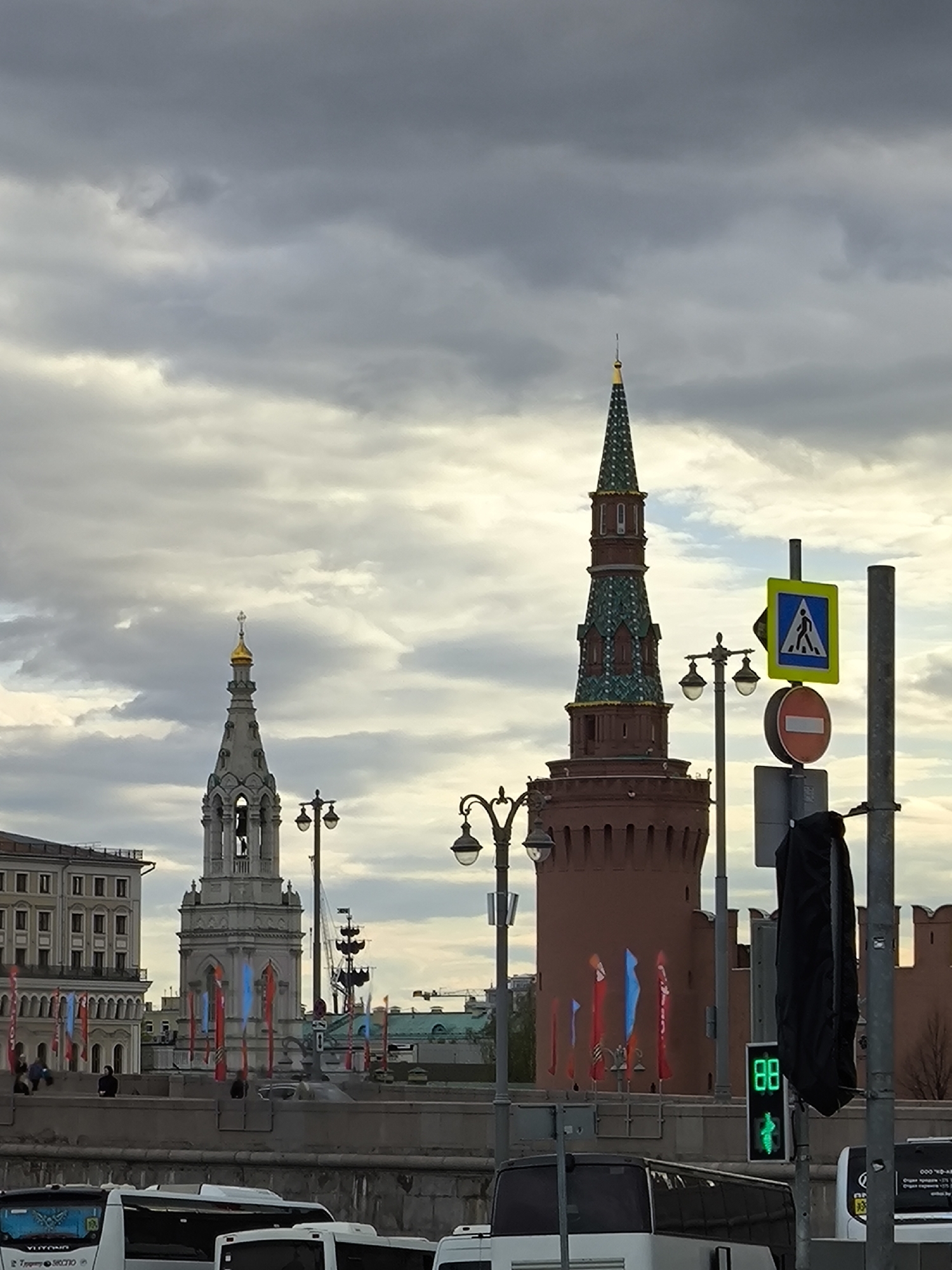 This is an eye-level, outdoor, color image taken in Moscow, Russia. In the background are two tall towers of different construction styles. The tower on the left is gray and topped with a gold dome. The tower on the right is brown with a green roof. Both towers are decorated with flags along the embankment. A few people are sitting on the stone wall, which extends the length of the image. In the foreground are streetlights and pedestrian crossing signals. White buses are in the lower portion of the image. Overcast skies fill the background.