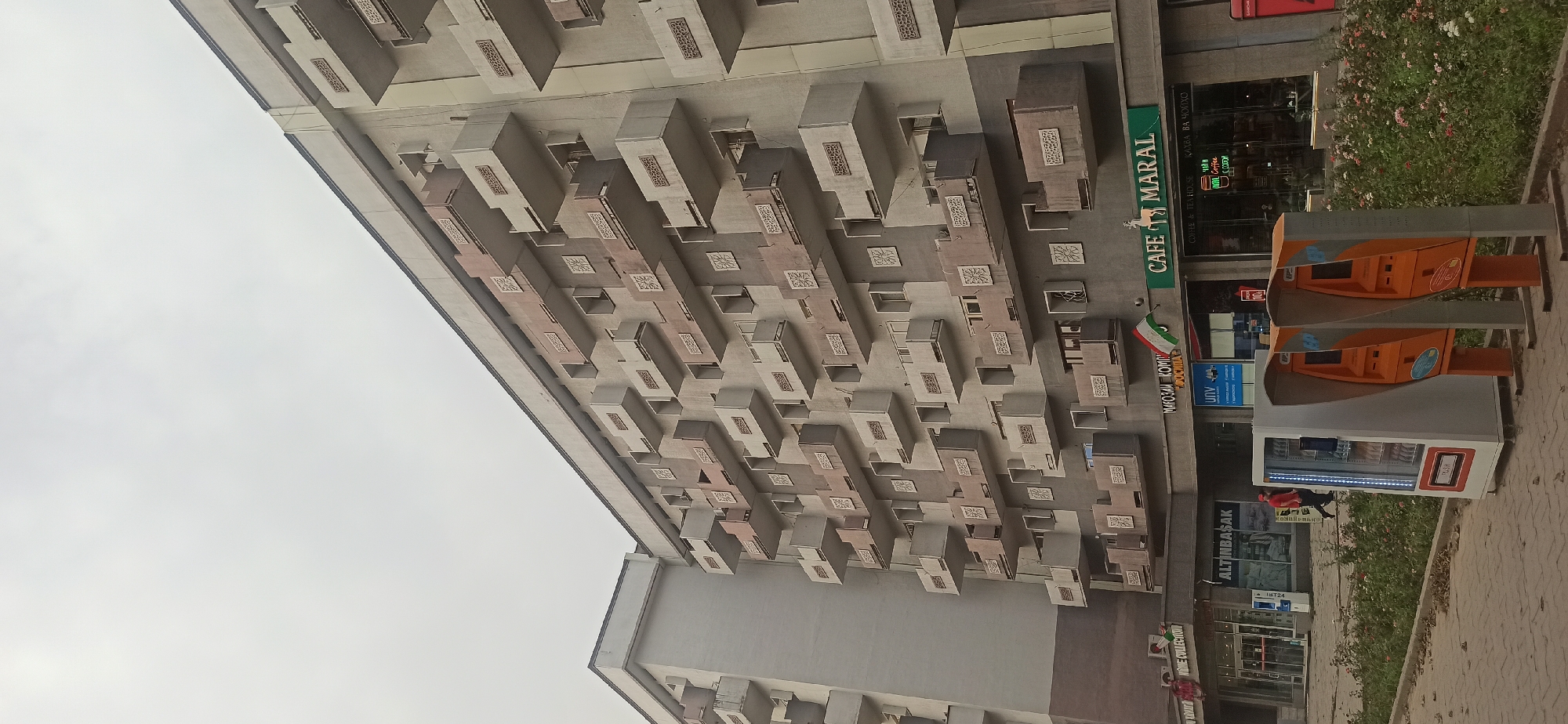 This image captures a street-level view of a multi-story apartment building with numerous balconies in Dushanbe, Tajikistan. The building has a gray facade with alternating cream and pink colored balconies, many featuring decorative square patterns. Below the building, a row of shops and cafes are visible. One prominent sign reads "CAFE MARAL" in green lettering, with a logo of a deer. Other businesses include "ALTINBASAK" and "HOME COLLECTION". To the left of the cafes, there is a white vending machine with a glass front displaying products, and to the right, two orange payment terminals are positioned on the sidewalk. A person in a red shirt and dark pants is walking past the vending machine. A flag, appearing to be the flag of Tajikistan, is mounted on the building above one of the shops. The sky is overcast, and the ground is paved with bricks. There is a small strip of greenery with flowering plants in front of the building and shops.