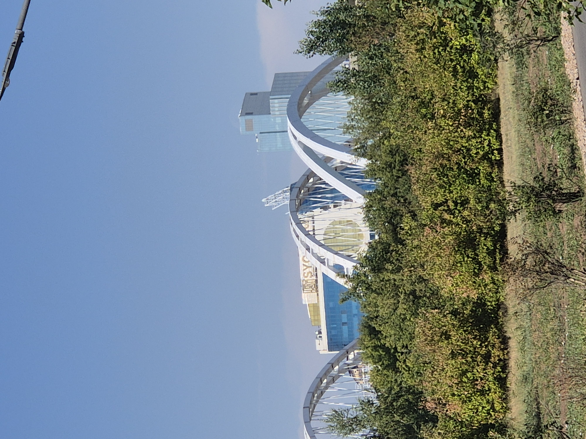 This image captures a wide shot of a modern cityscape in Astana, Kazakhstan, dominated by a clear blue sky and lush green trees in the foreground. In the mid-ground, several white, arching bridge structures are visible, indicative of a significant urban infrastructure project. Behind the bridges, modern buildings rise, featuring glass facades that reflect the sunlight. One prominent building displays the "SYC" logo in gold lettering. The environment appears to be an outdoor public space, likely a park or a vantage point offering views of the city's development.