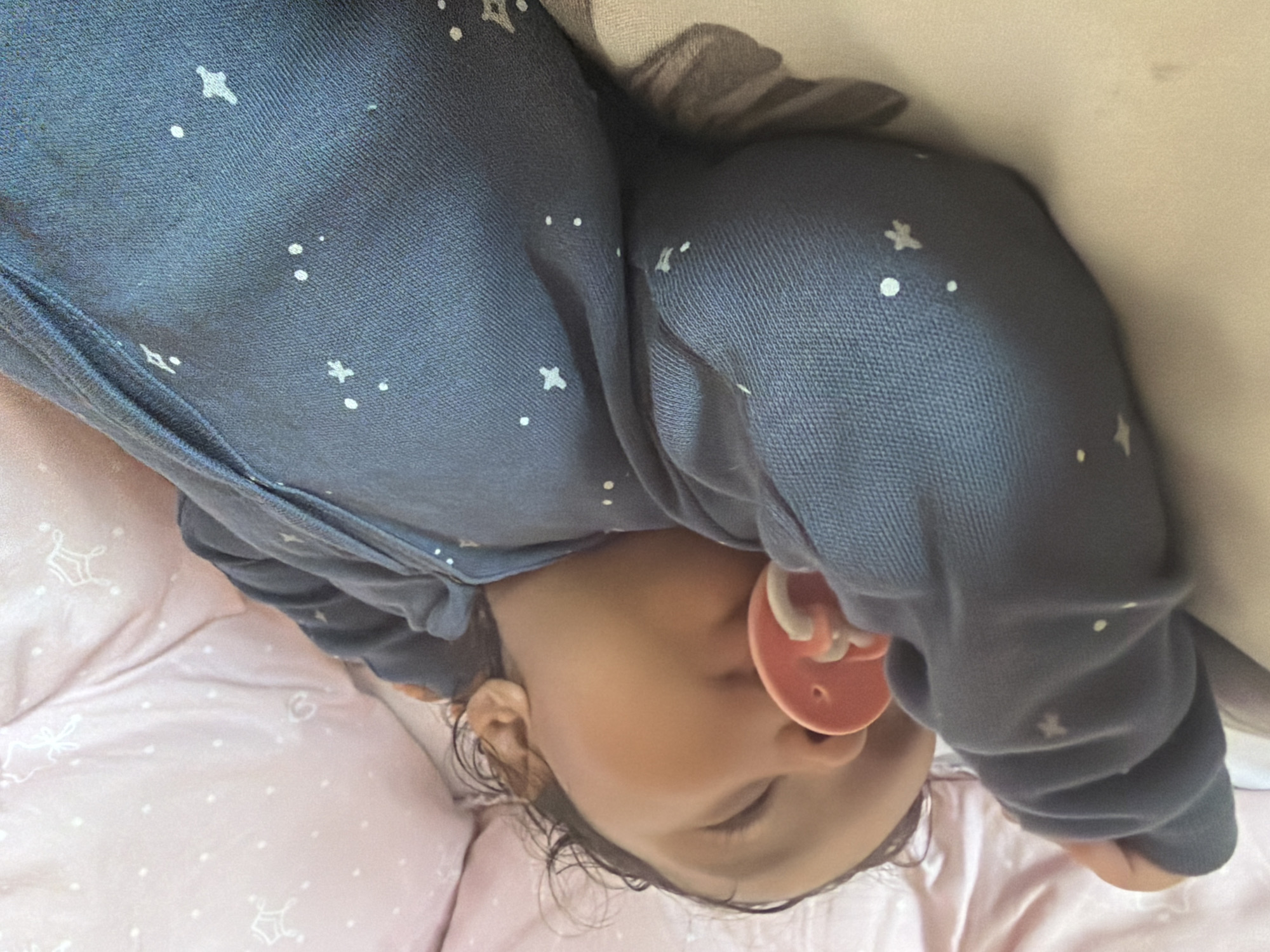 A dark-skinned baby sleeps on a light-colored surface, propped up on their left arm. The baby is wearing a dark blue onesie with a pattern of white stars. A pink pacifier is in the baby's mouth. The background consists of light pink fabric with a similar star pattern. The scene appears to be a domestic setting, possibly a bed or crib, located in Razhevo Konare, Bulgaria.