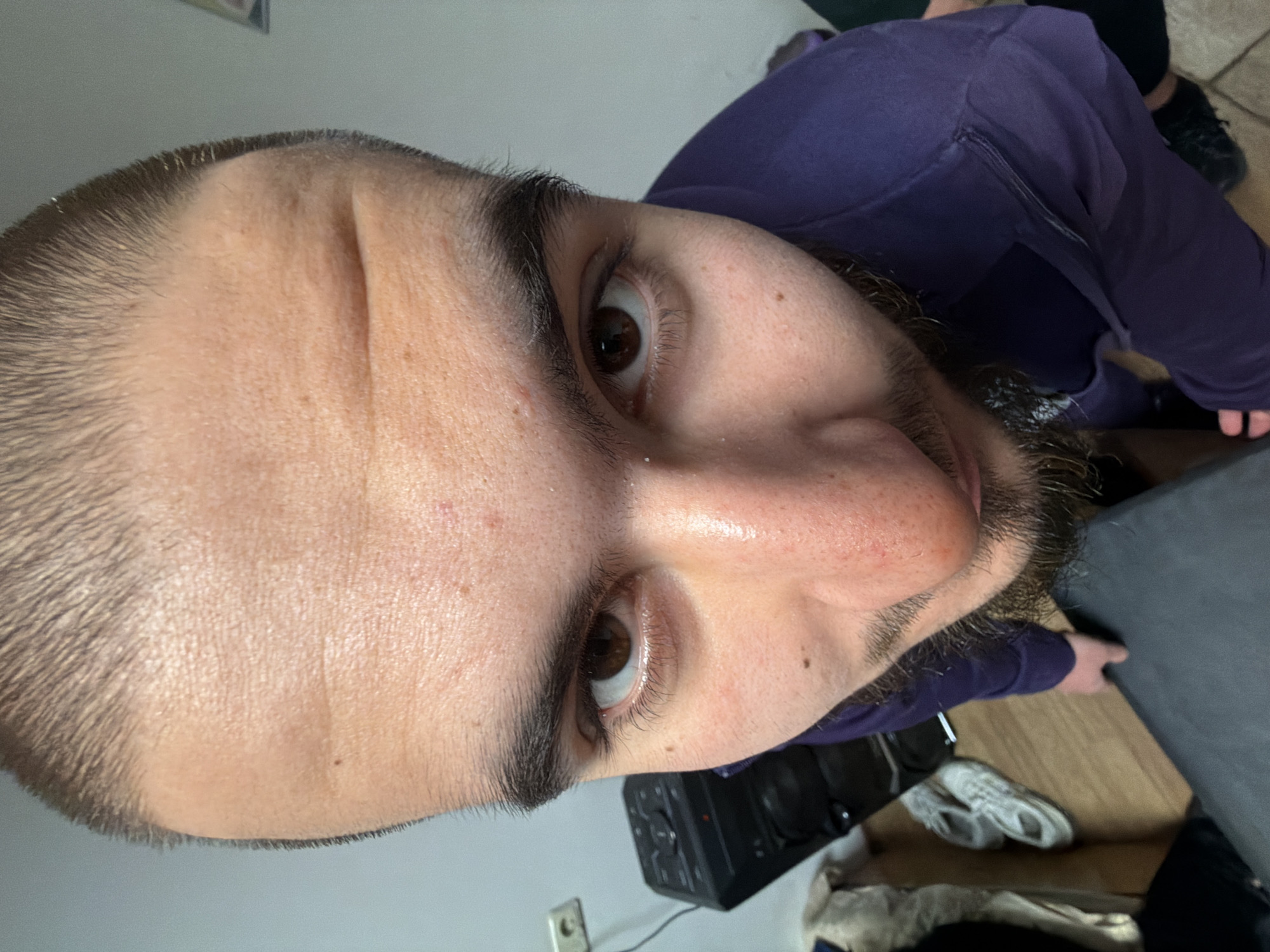 A close-up, distorted perspective photograph shows a person's face angled upwards. The individual has short, dark hair, brown eyes, and a beard with visible stubble. The skin shows pores and minor blemishes. The person is wearing a purple, long-sleeved garment. The background includes a white wall, a black object (possibly a speaker), a power outlet, a wooden floor, a pair of white sneakers, and a dark grey surface. There is no indication of a specific location in Plovdiv, Bulgaria, or any other location.