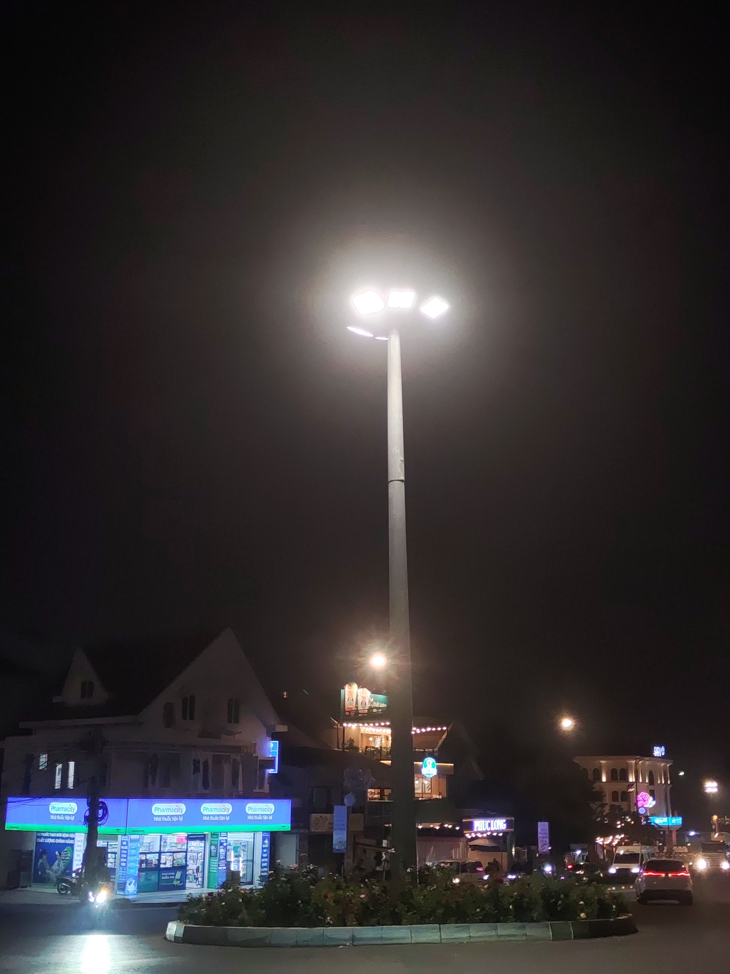 The image depicts a night scene in Da Lat, Vietnam. Prominently featured is a tall, central light pole with multiple bright lamps illuminating a roundabout. The road is busy with both vehicular and pedestrian traffic. A motorcycle moves past a series of Pharmacity drug stores located under a two-story building with a pointed roof design. Further in the background are other lit buildings, including one with the sign "PHUC LONG." The overall impression is of an active urban area at night.