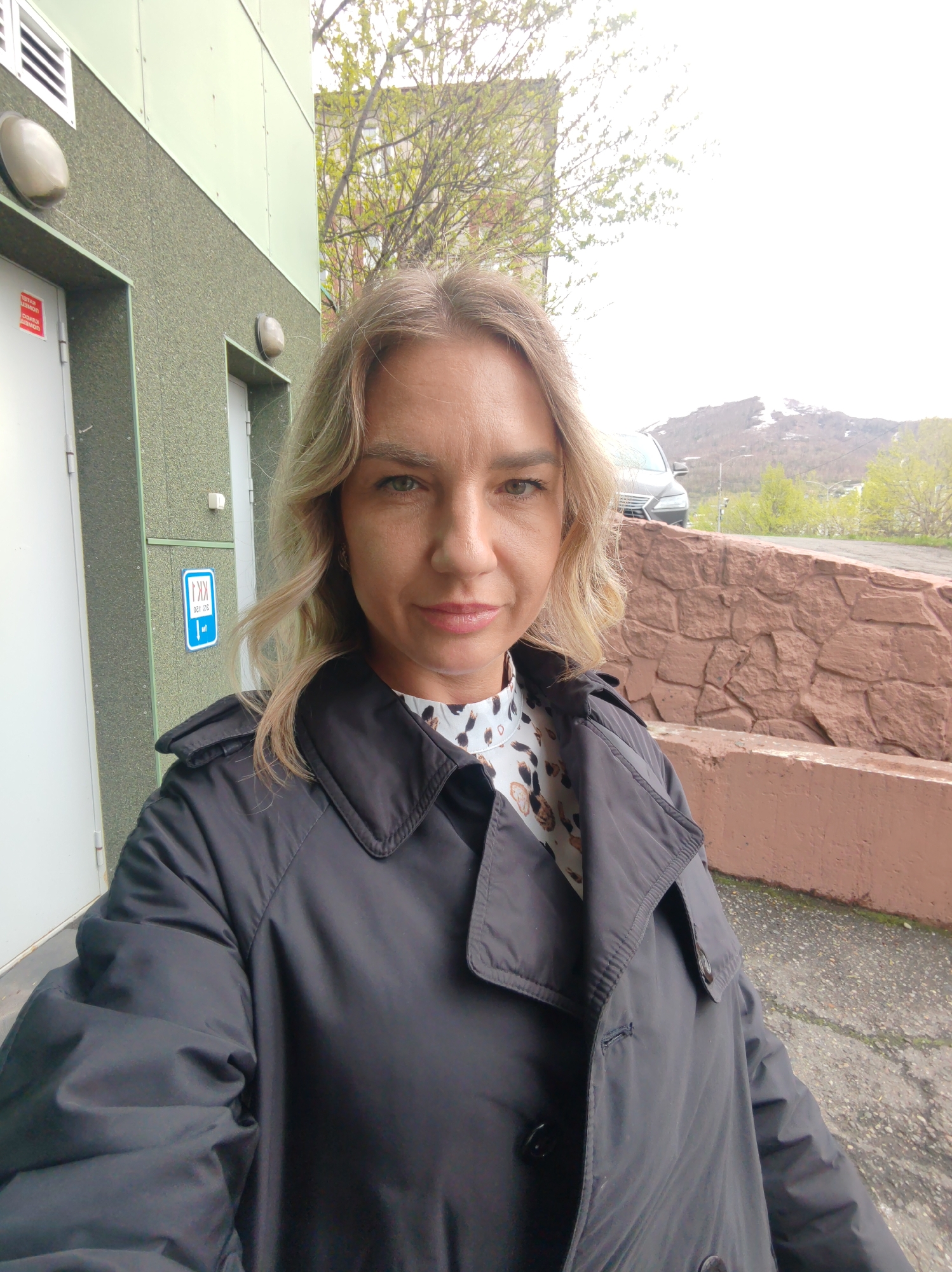 A woman is standing outside near a building with a green and white facade. The woman has blond hair and is wearing a black jacket over a patterned blouse. Behind her is a wall with a red brick texture and a car parked on a road. In the background, a mountain range can be seen. The location is likely Petropavlovsk-Kamchatsky, Russia.