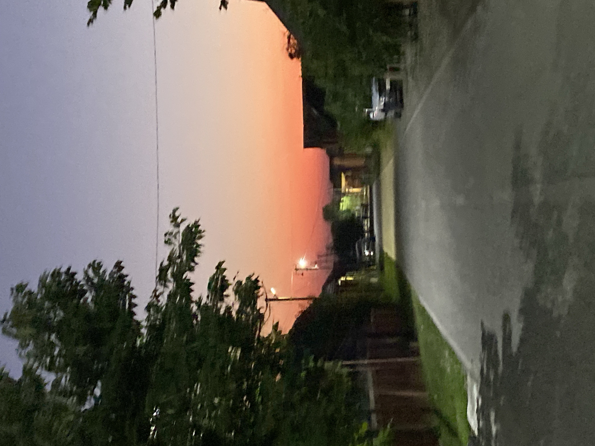 The image shows a street scene at dusk in Alkhan-Kala, Russia. A paved road leads into the distance, flanked by trees and houses on either side. The sky is a gradient of purple and orange, indicating the sunset. There are streetlights visible, casting pools of light on the road. Two cars are parked on the right side of the road. The overall atmosphere is peaceful and quiet, typical of a residential area at this time of day.