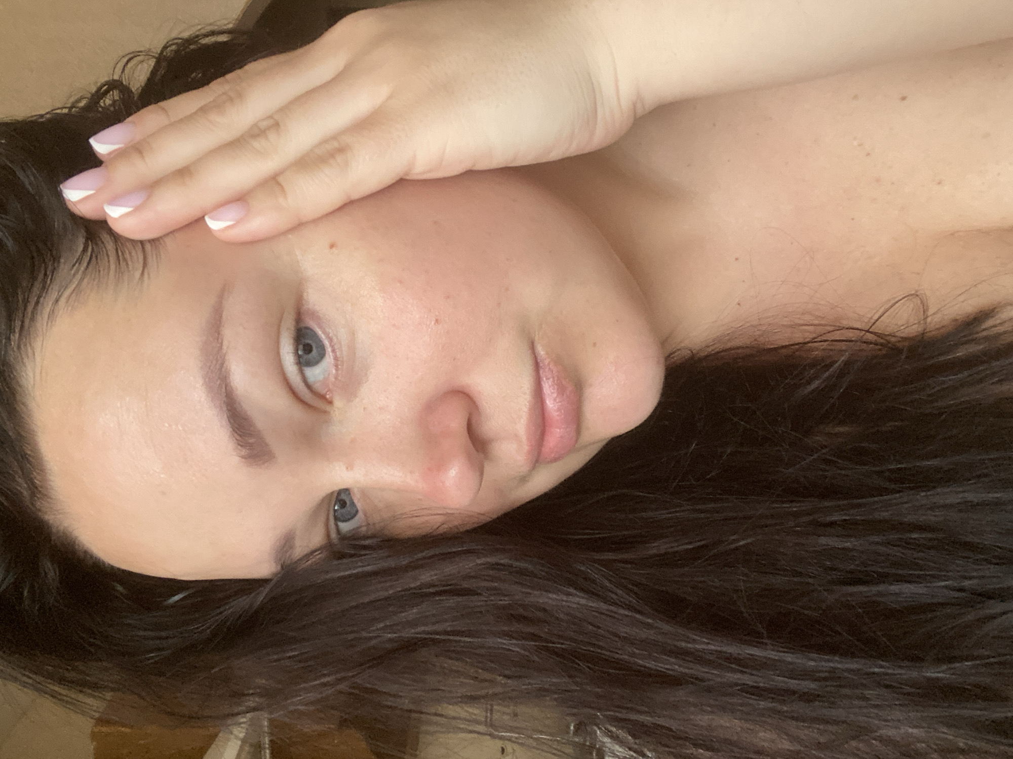 A young woman with fair skin, dark hair, and blue eyes is lying down, facing the camera. Her right hand is placed on her forehead. She has a French manicure. The background is blurry and indistinct, revealing little about her location. There is no evidence to suggest she is located in the city of Sochi, Russia.