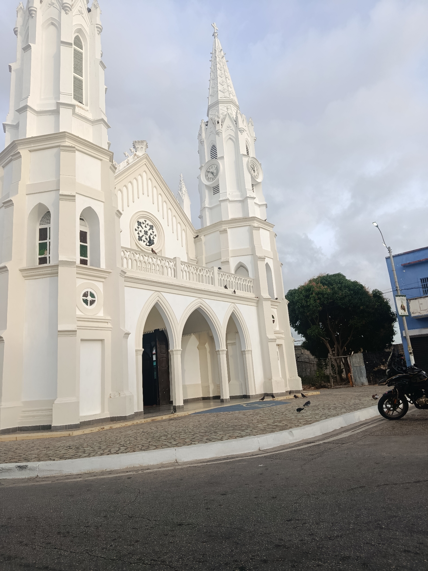 The image shows the Basilica Menor de Nuestra Señora del Valle, a prominent white church in Juan Griego, Venezuela. The church has two towers, one with a clock face, and multiple arched entrances. The sky is overcast. The street in front of the church is paved, with a motorcycle parked on the edge. Several pigeons are on the pavement near the church entrance. A tree and a blue building are visible in the background.