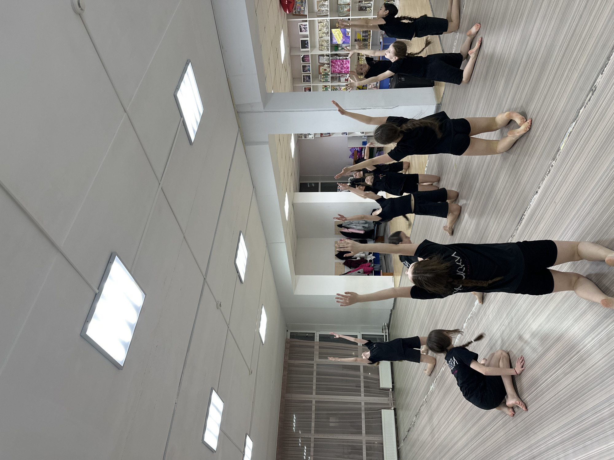 A group of girls are in a dance studio in Votkinsk, Russia. They are all wearing black tops and shorts or skirts. Some of the girls are standing and stretching, while others are sitting on the floor. The studio has a tiled floor and white walls. There are windows along one wall and a mirror on another. There are rectangular ceiling lights. Shelves filled with items line part of one wall.