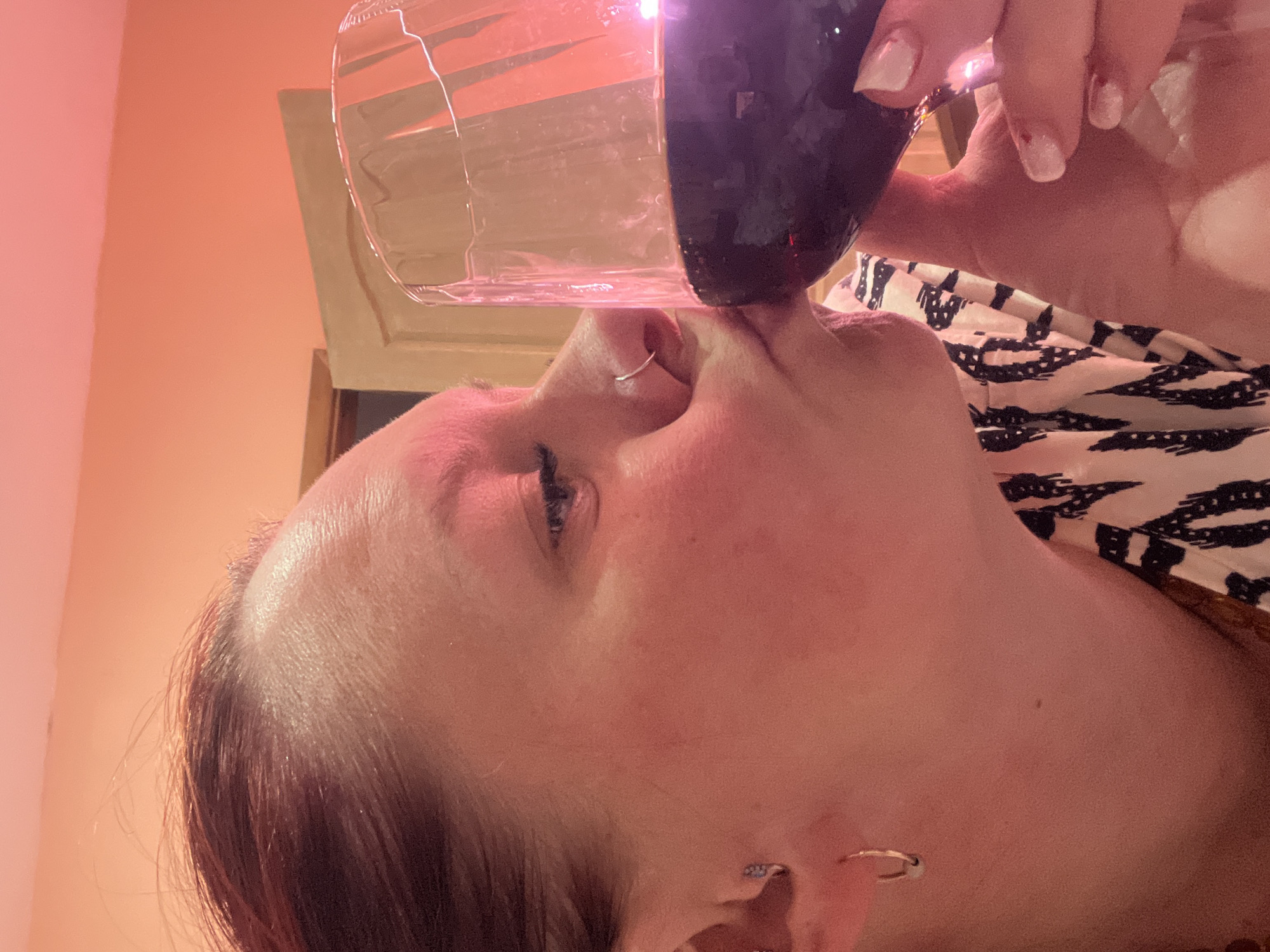 A woman with red hair is drinking from a wine glass filled with a dark liquid. The woman is wearing a black and white patterned shirt. She has a ring in her nose and an earring. Her fingernails are painted with white tips. The environment suggests an indoor setting with warm lighting. There is no information available to determine if the location is Chaikovsky, Russia.