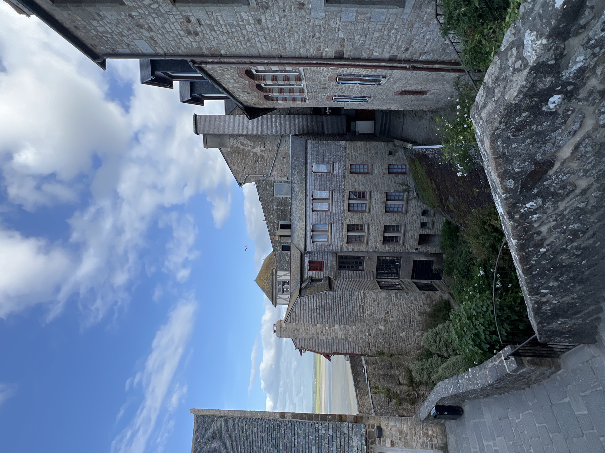 This image depicts a scenic view within the fortified commune of Le Mont-Saint-Michel, France, captured on a clear day with a blue sky and scattered white clouds. The scene is dominated by historic stone buildings with varying architectural details, nestled together on a steep incline.
In the foreground, a stone staircase leads downwards to the left, bordered by a rough-hewn stone wall topped with dark lichen. A dark trash bin is positioned next to the wall on the left. Over the wall and descending further, a dark metal railing partially frames an area of dense green foliage, including bushes and plants with small white flowers.
The mid-ground features a complex arrangement of buildings constructed primarily from grey stone blocks. Roofs vary, with some being slate and others appearing to be clad in wood shingles. Multiple windows with dark frames are visible, some accented with red trim. Several prominent stone chimneys rise above the rooftops. A narrow, dark stone alleyway or passage winds between the buildings, descending further into the scene. One building on the far right displays arched windows with reddish-brown brickwork and a vertical reddish-brown pipe running down its facade.
In the background, beyond the cluster of buildings and their surrounding greenery, a broad, flat expanse of light brown and green land is visible, characteristic of an estuary at low tide. This suggests the proximity to the surrounding bay. A small, dark bird is silhouetted against the blue sky in the upper right quadrant of the image. No people are present in the scene.