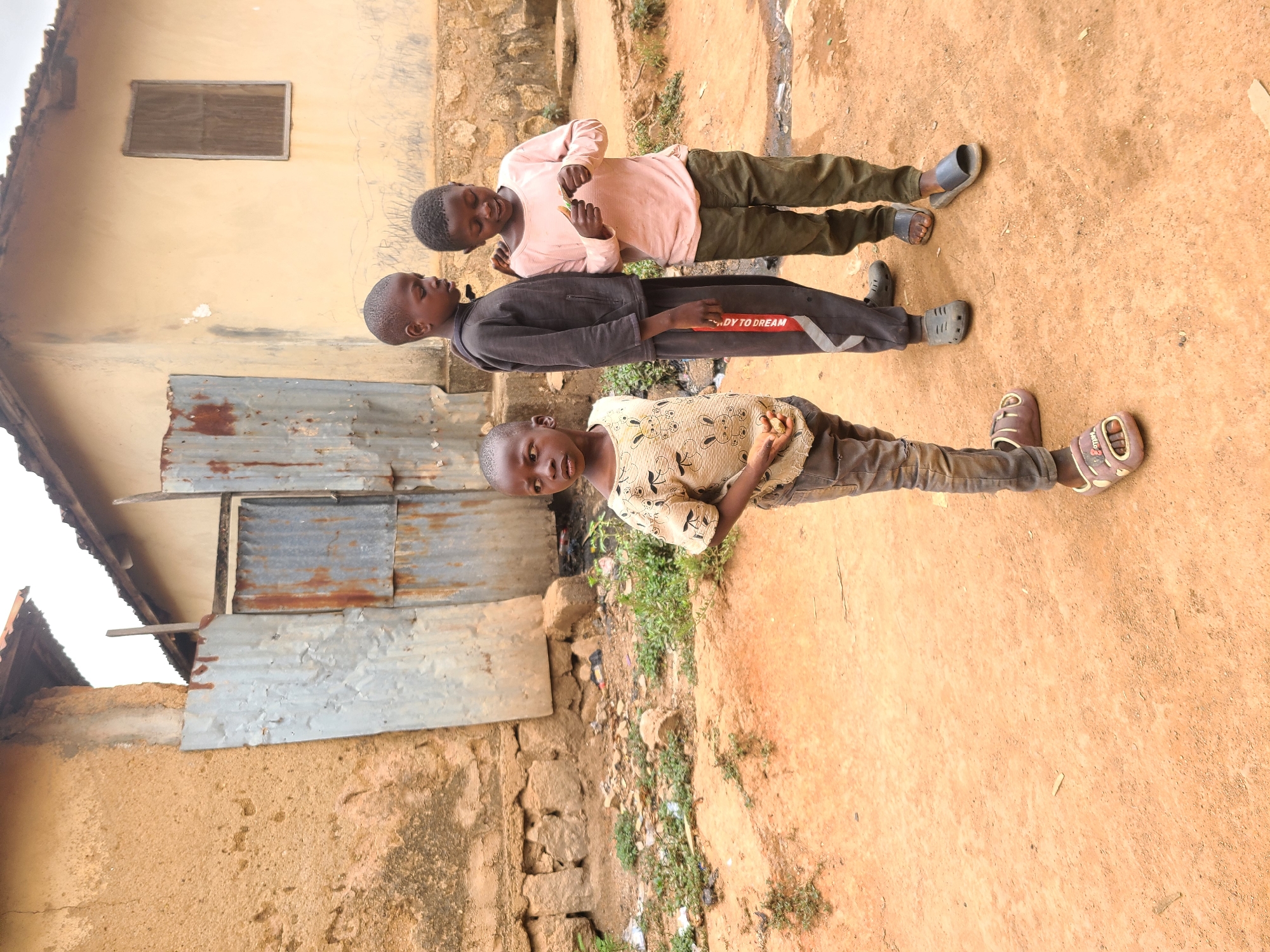 Three children are standing in a dirt yard next to a dilapidated building. The building has a combination of mud brick, stone, and corrugated metal siding. One window is visible on the upper part of the building. The ground is reddish-brown dirt. The child closest to the viewer is wearing a patterned shirt and brown pants. The child in the middle is wearing a dark sweatshirt and pants. The child furthest from the viewer is wearing a pink shirt and green pants. The environment suggests a lower-income neighborhood. The location is likely Jos, Nigeria, based on the context and the appearance of the buildings and landscape.