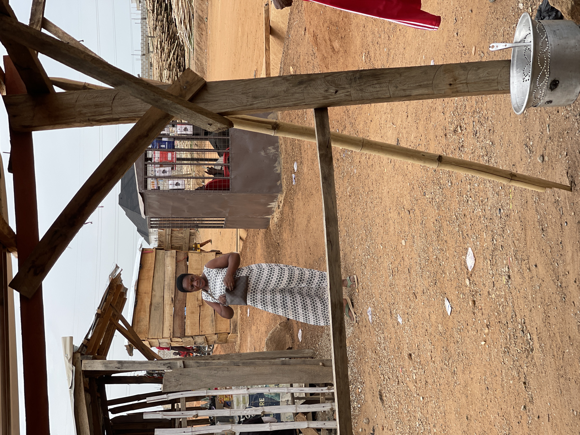 The scene is in Angwan Sayawa, Nigeria. The ground is dry, red dirt with scattered debris. A woman in a patterned jumpsuit stands in the middle ground, holding a dark purse. She is wearing sandals. Behind her is a small shop constructed from wooden planks and corrugated metal. The shop displays boxes of merchandise behind a metal cage. To the left, a partially constructed wooden structure frames the view. Power lines are visible in the distance. An aluminum pot is attached to a wooden pole. Stacks of bamboo sticks are visible in the background.